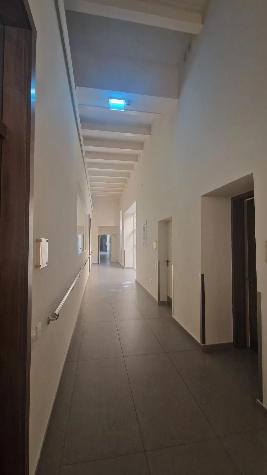 Long corridor in a modern building with white walls, grey tiled flooring, and large windows on the left side. There is a handrail along the wall, and ceiling lights providing illumination. Doors are visible on the right side of the corridor.