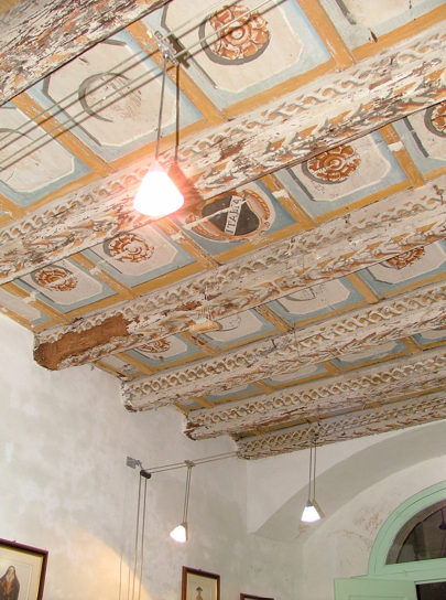 Decorative wooden ceiling with ornate painted and carved details in a historic interior, with hanging light fixtures.