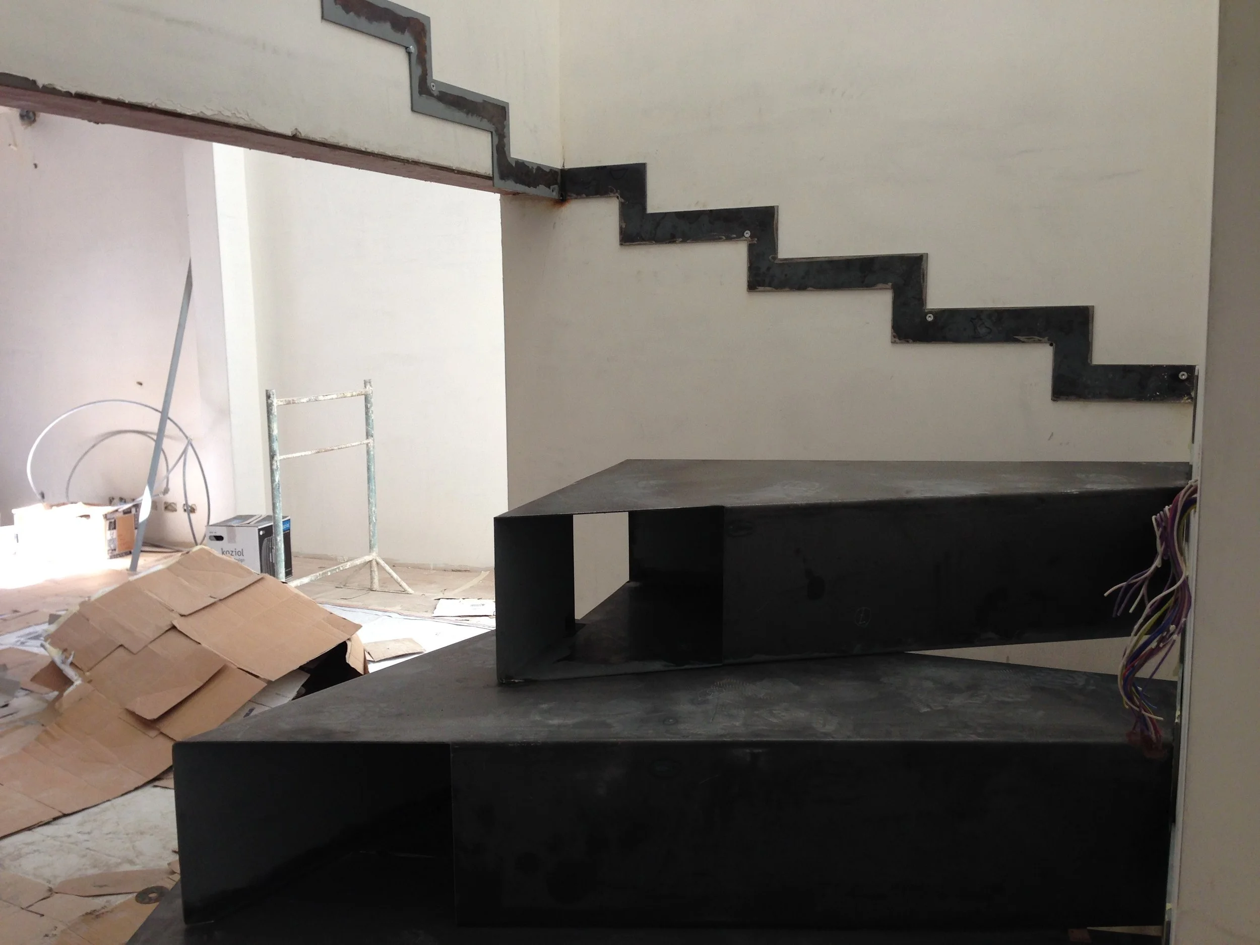 Interior of a building under construction, showing a staircase framework with metal framing, unfinished walls, and construction materials scattered on the floor.