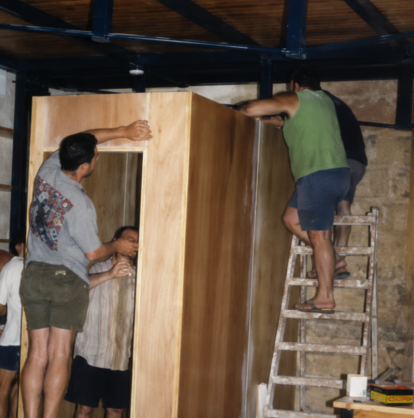 Men working together to assemble a large wooden structure inside a building, with one man standing on a ladder and others supporting and guiding the piece.
