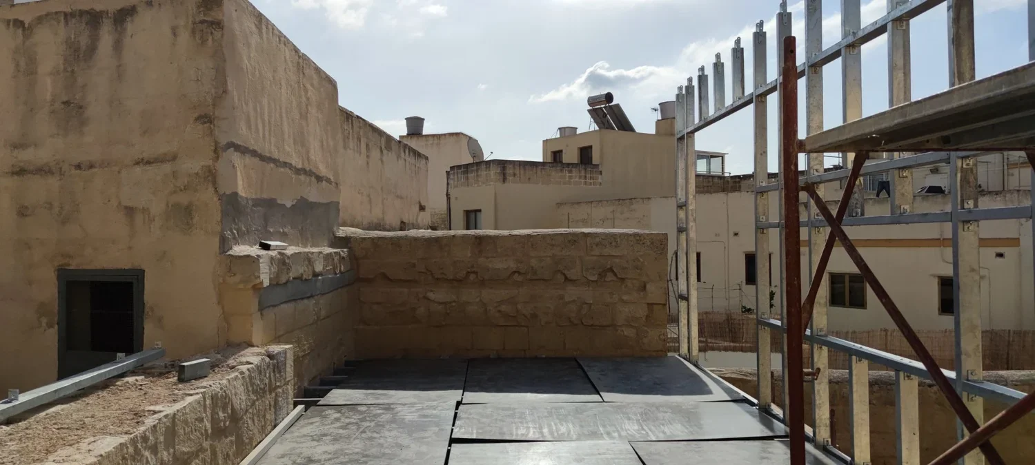 View of a construction site on a rooftop with scaffolding on the right, stone walls and buildings in the background, partly cloudy sky.