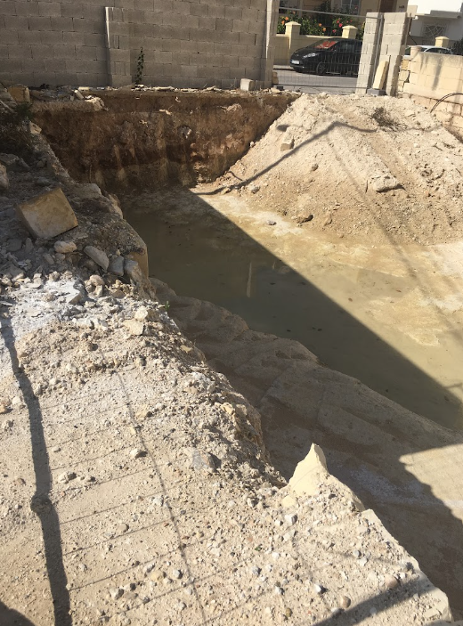 A dug-out construction site with exposed dirt and water, surrounded by concrete blocks and a partially built wall, with a street and parked cars visible in the background.