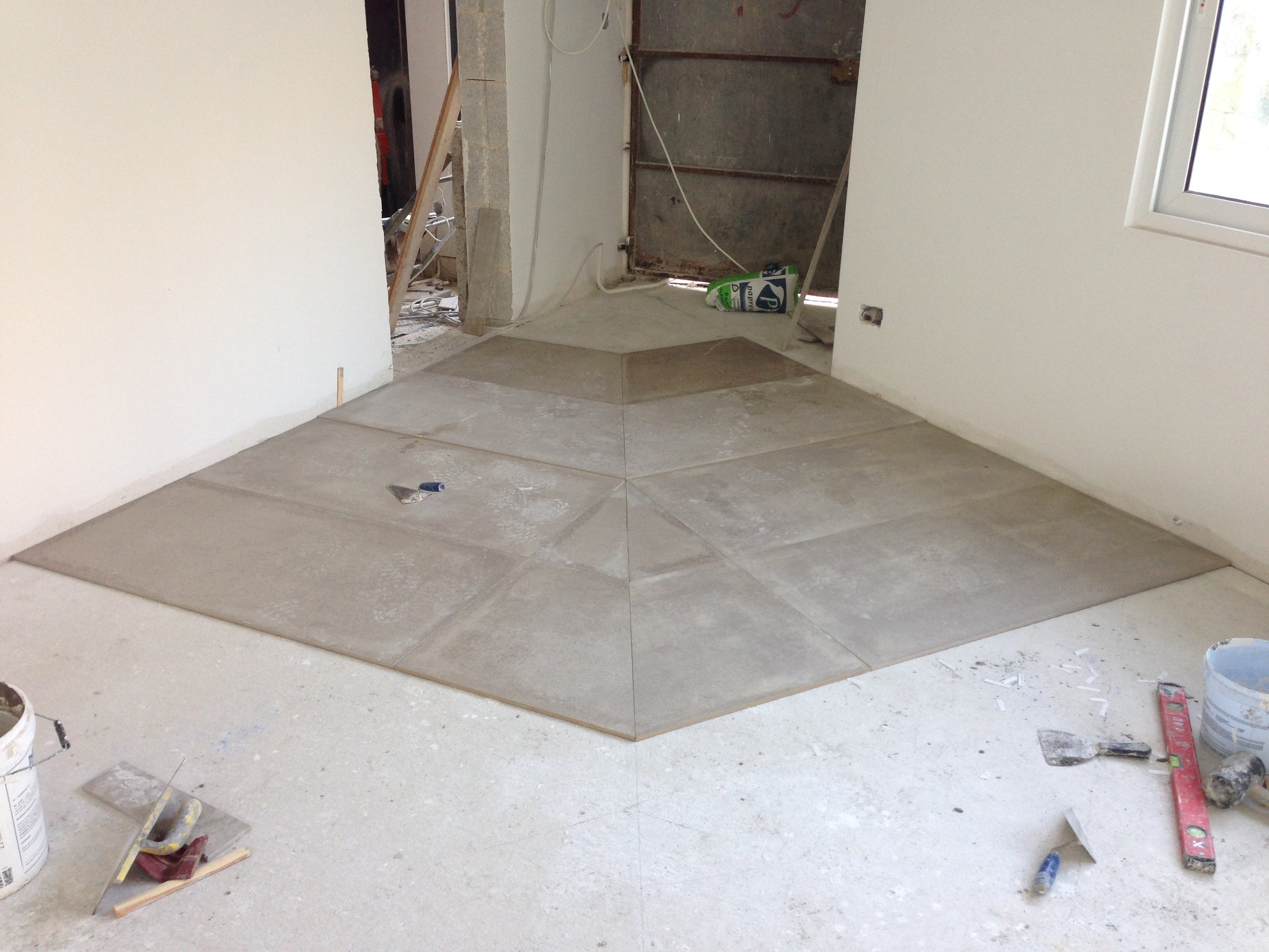 Interior of a room under construction with tiled flooring and construction materials scattered around, leading to a partially open door revealing an unfinished space.