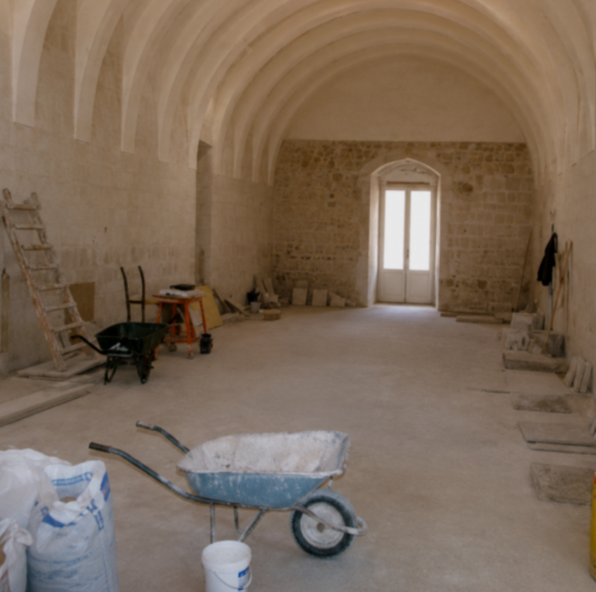 Interior of a room under construction with stone walls and arched ceiling, construction tools and materials including a wheelbarrow, ladder, and buckets present.