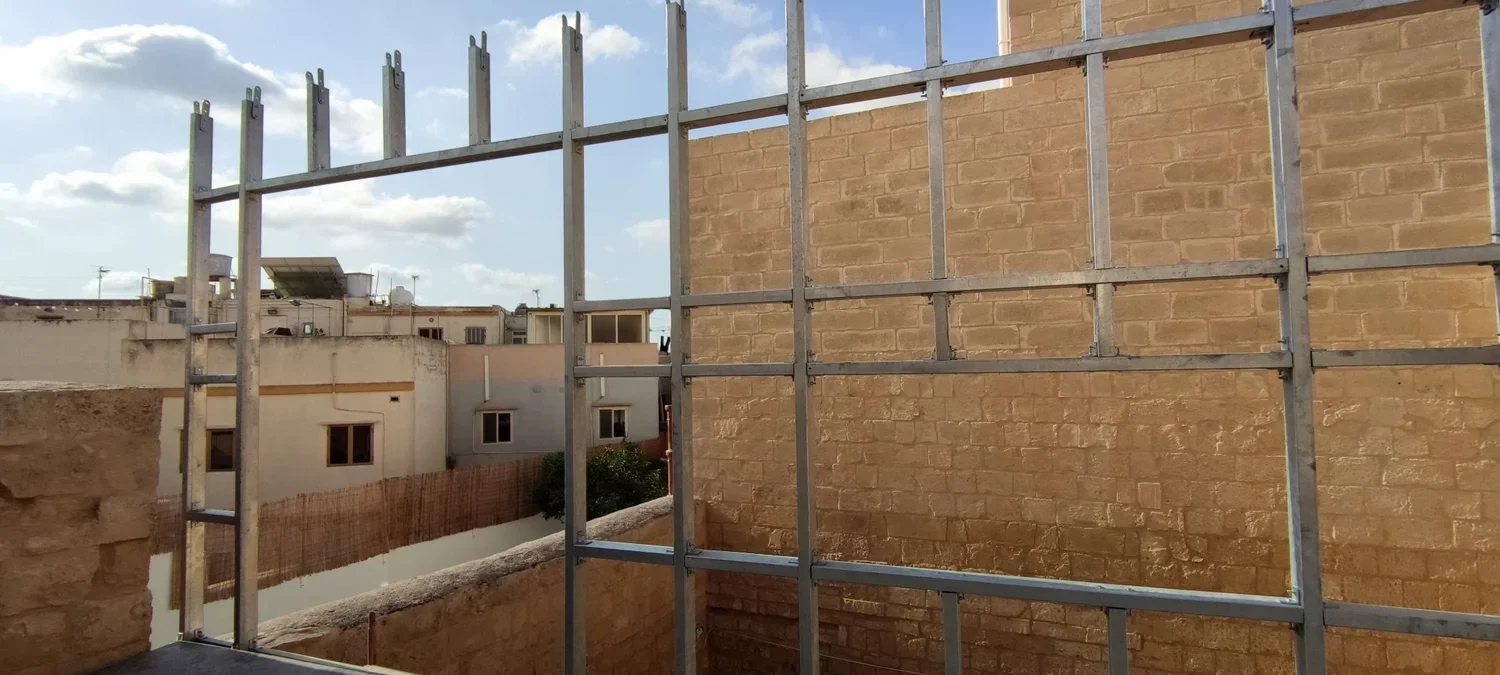 View of a construction site with a metal framework for a building and brick walls, with neighboring buildings and a partly cloudy sky in the background.