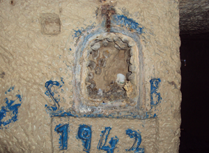 Close-up of an old stone wall with a small, arched niche containing a weathered cross and some shells. Blue and white paint markings, including the year 1962, are visible on the wall.
