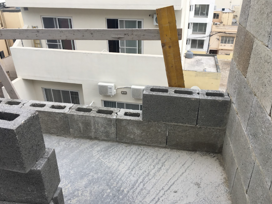 View from a building under construction showing cinder blocks forming a low balcony wall, with wooden beams and a plank, overlooking neighboring apartment buildings with air conditioning units and windows.