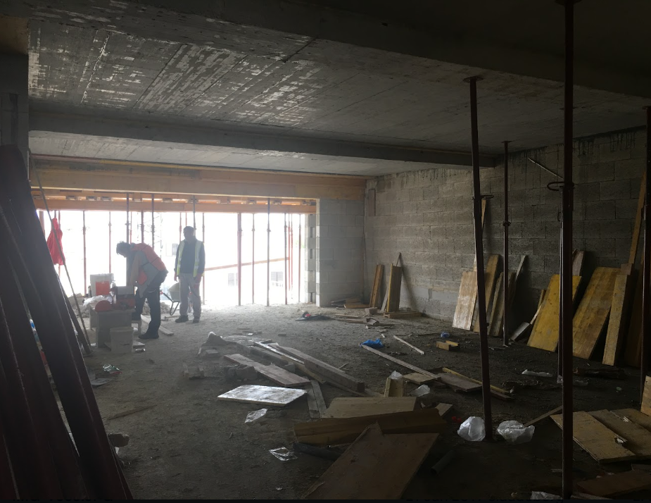 Construction site with two workers inside a partially built building, surrounded by scattered wooden planks, construction materials, and support poles, with sunlight coming through an open wall.