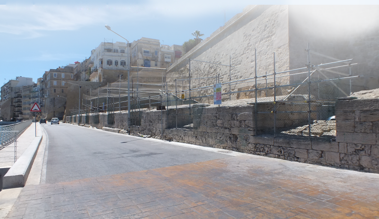 Street view with stone wall, scaffolding, and buildings on a hillside in a sunny area.