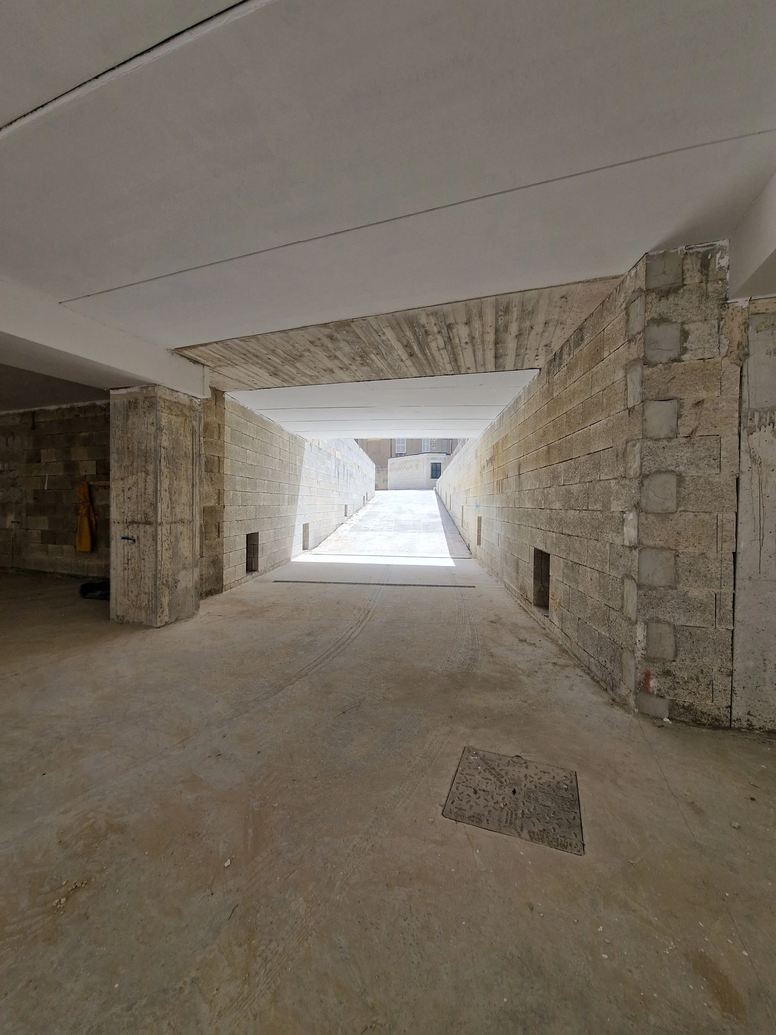 An underground passageway with unfinished construction, showing concrete and brick walls, a concrete arch, and a bright exit in the background.