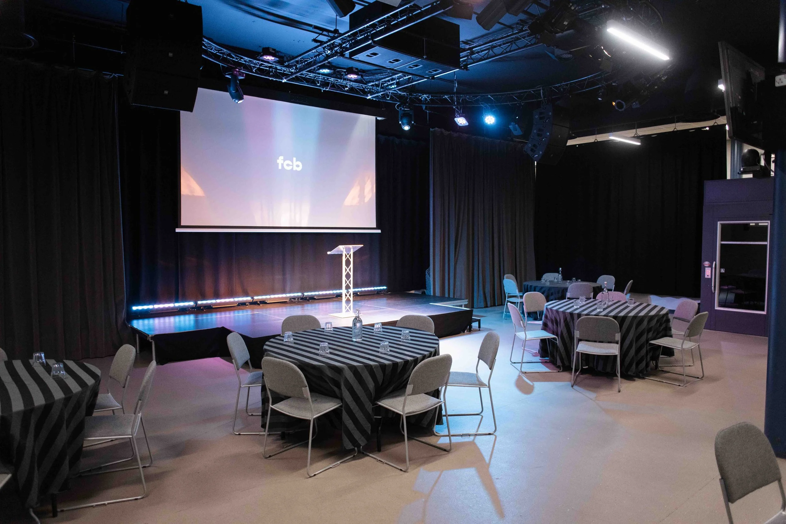 An indoor event venue with round tables covered in black and white striped tablecloths, set with glassware and chairs. There is a stage with a podium, a large projection screen and overhead stage lighting. The room has black curtains and a ceiling ri