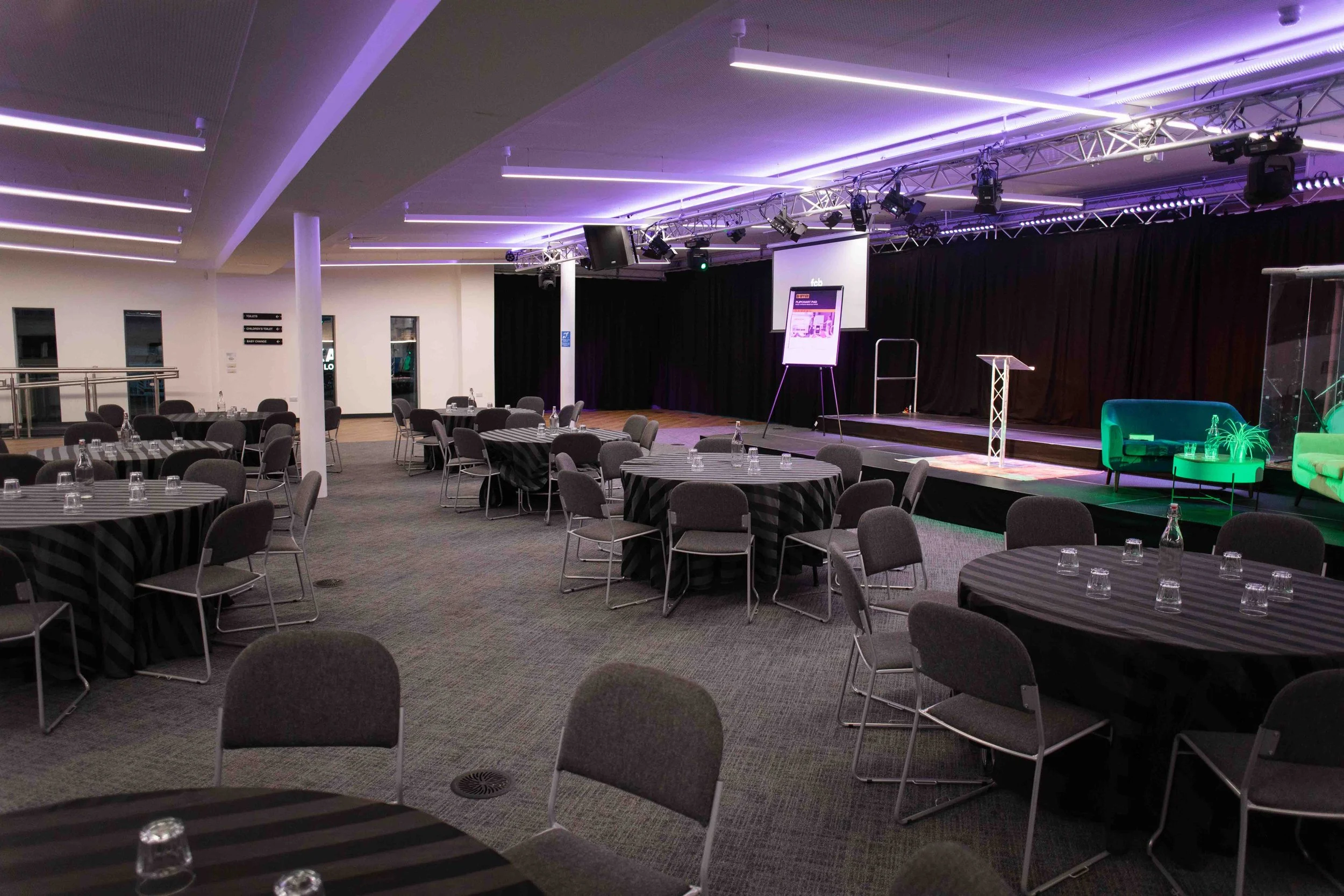 Bradford event space with round tables covered with black and gray striped tablecloths, glass water bottles, and glasses. A stage with black curtains, a large screen, and lighting equipment is set up at the front.