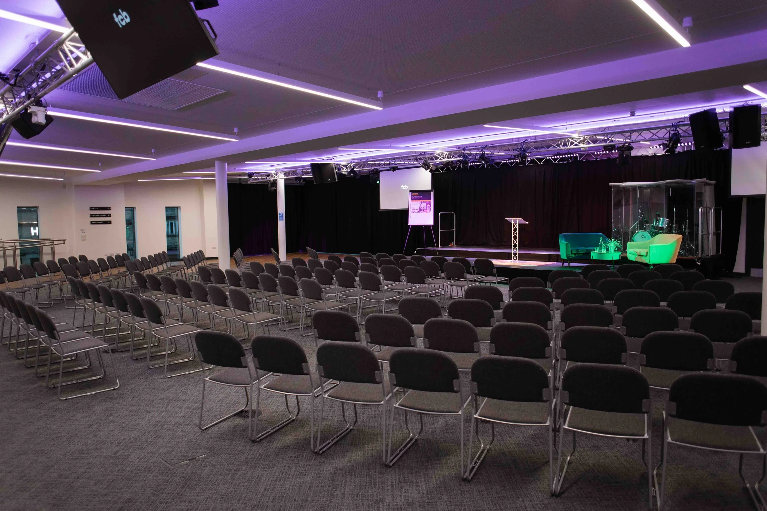 A conference venue in Bradford with rows of black chairs facing a stage with a podium, musical instruments, and large screens. Purple and white ceiling lights illuminate the room.