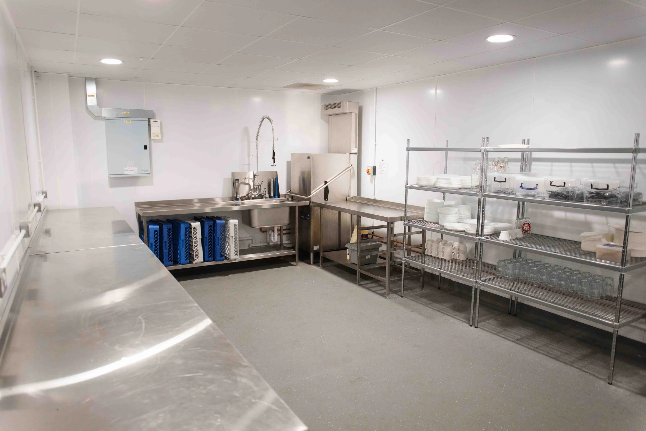 Commercial or industrial kitchen with stainless steel countertops, a sink, shelves with dishes and glasses, and white walls.