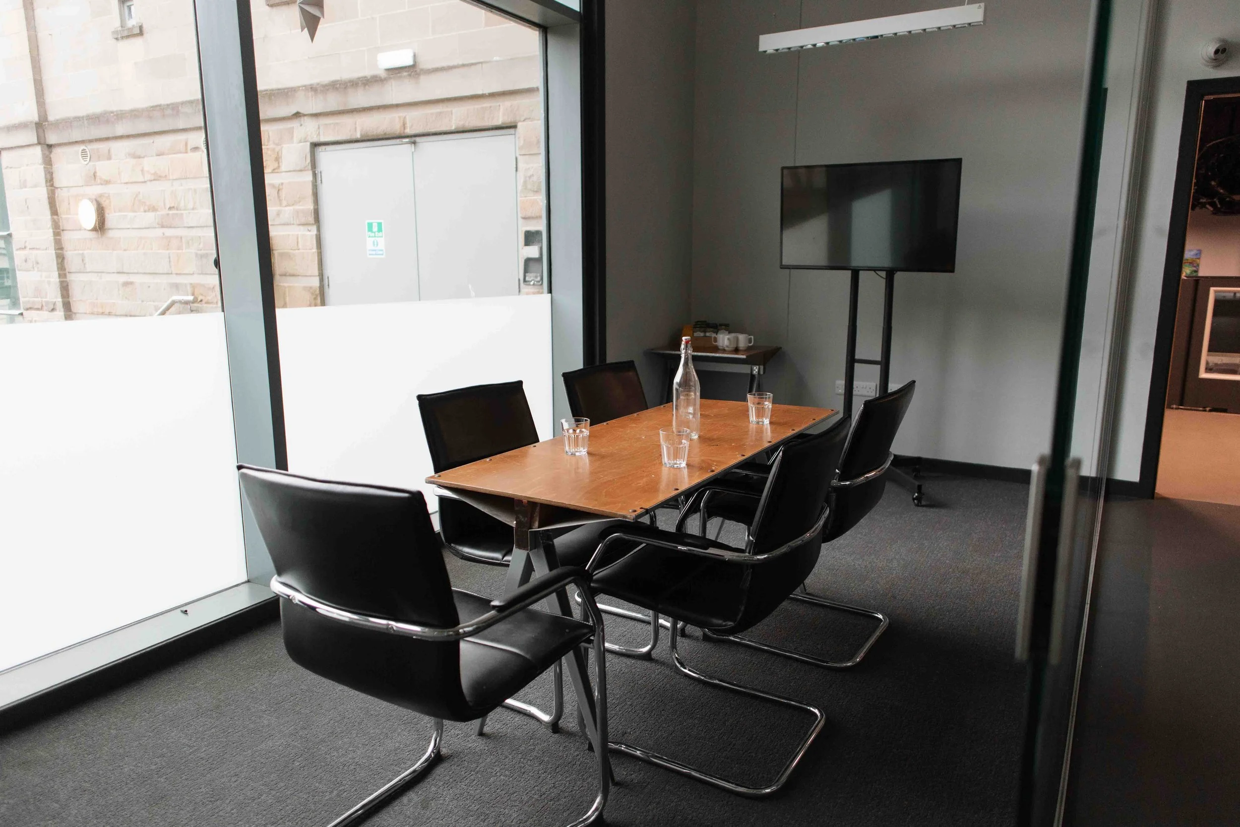 A Bradford conference room with a wooden table surrounded by six black chairs, a glass bottle of water, and four glasses on the table. There is a small table with coffee mugs and supplies, and a window showing an exterior brick building.