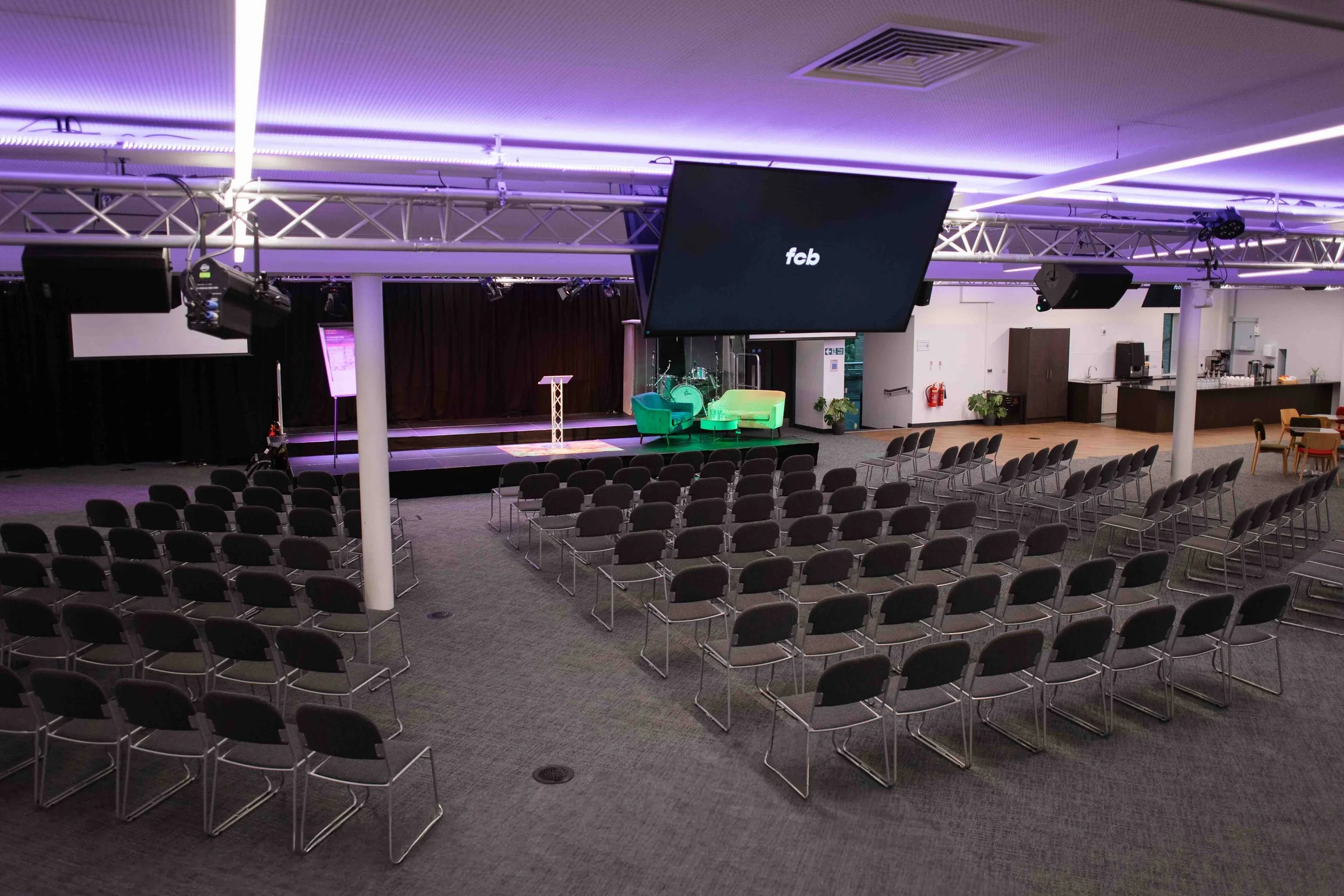 Conference room for hire in Bradford with rows of chairs, a stage with green chairs, large screen displaying 'fcb', and purple lighting.