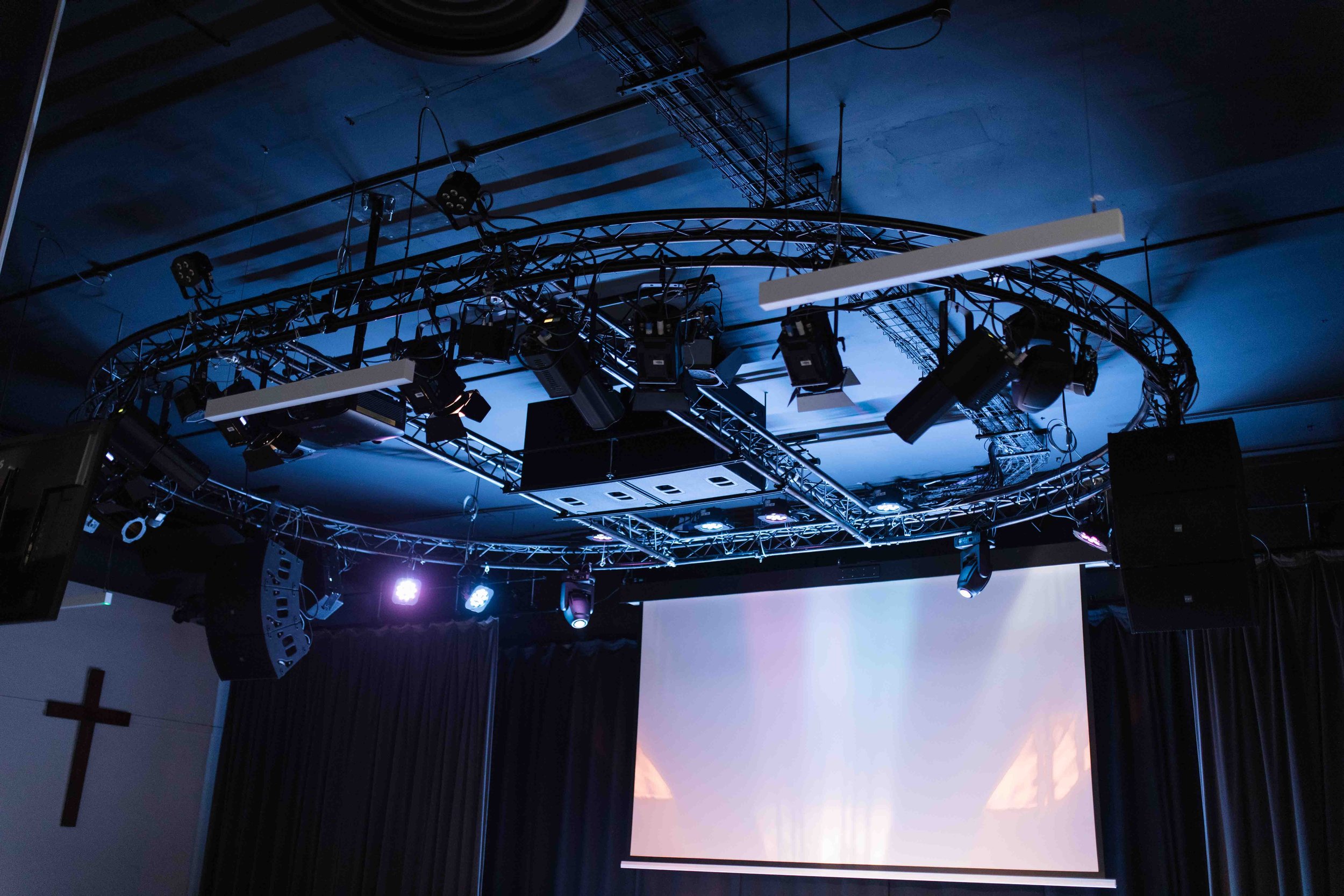 A stage with mounted lighting fixtures, a large projection screen, and black curtains, suggesting a setup for a presentation or event.