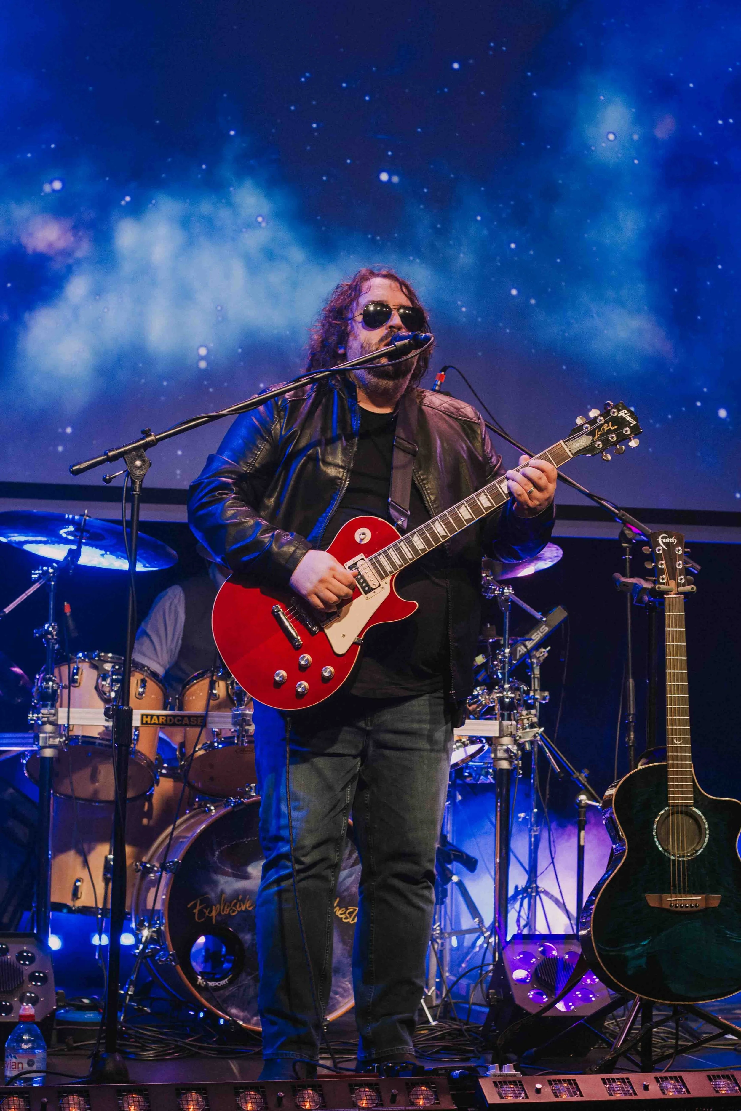 Musician with dark curly hair and sunglasses playing a red electric guitar on stage with a drummer in the background, and outer space-themed backdrop.