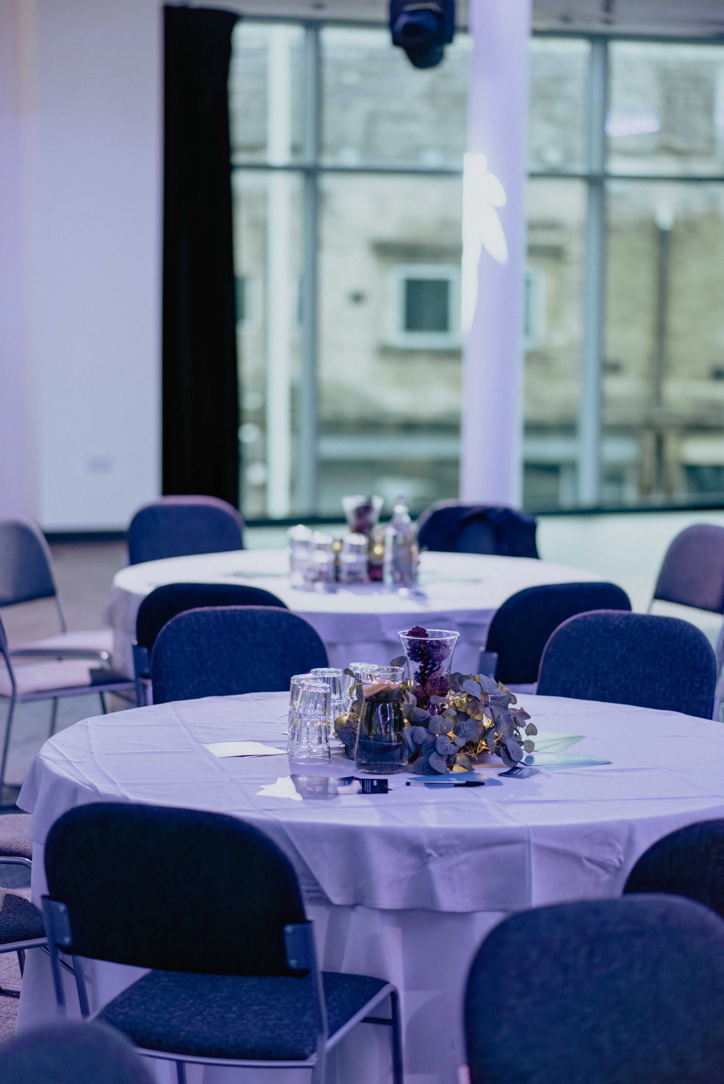 A conference or event room with round tables covered with white tablecloths, decorated with small flower arrangements, glasses, and bottles. Chairs are arranged around the tables. Large windows in the background let in natural light.