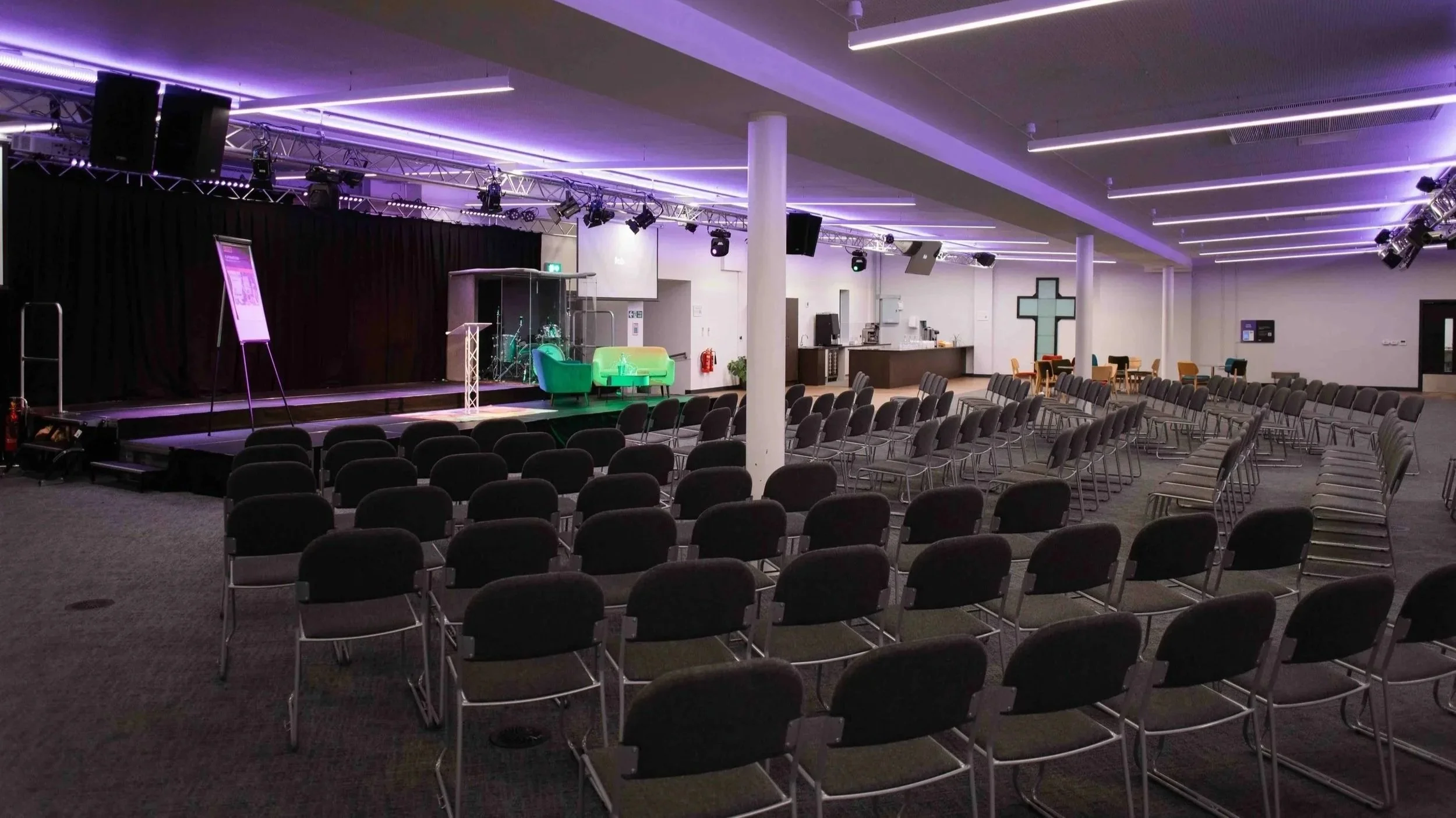 Large conference room for hire in Bradford with rows of empty chairs facing a stage with musical instruments and lighting equipment, purple and white neon ceiling lights, and a cross on the wall.