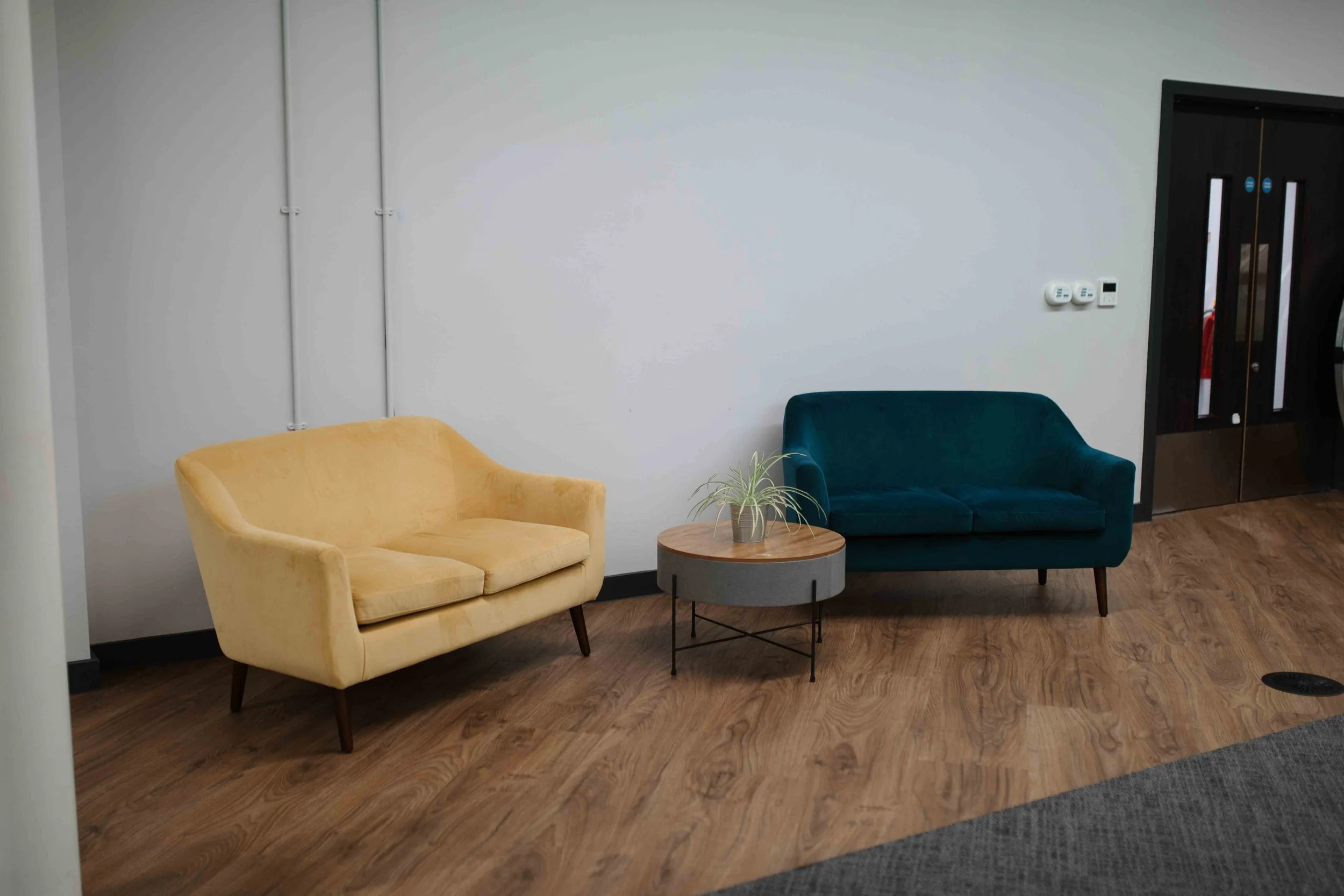 A yellow armchair and a teal sofa are arranged facing each other in a room with white walls and wooden flooring. A round wooden coffee table with a small plant is placed between the chairs. A black door with glass panels and a set of white thermostat