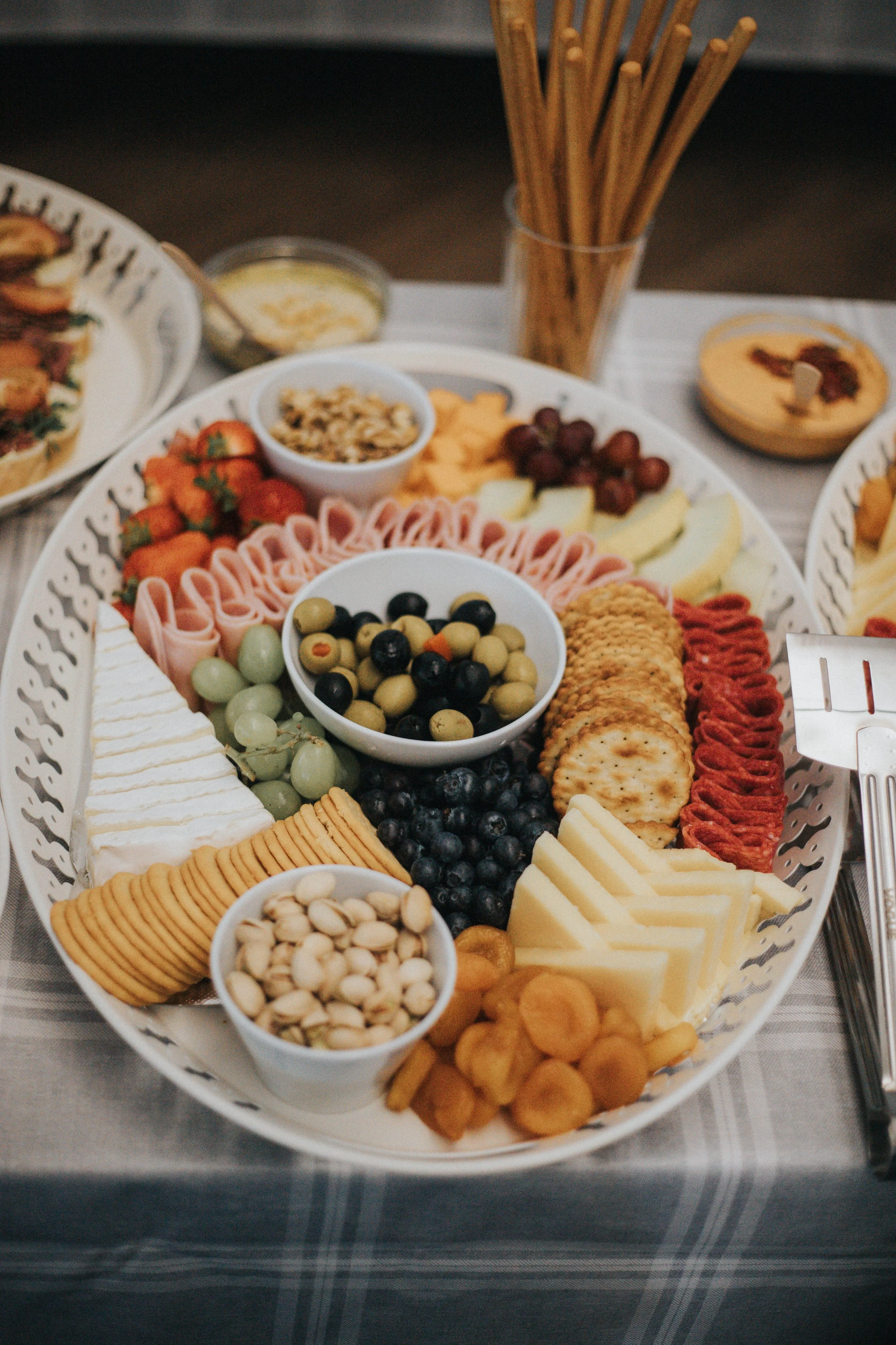 A large white platter with assorted cheeses, crackers, grapes, strawberries, olives, nuts, and other snacks, accompanied by small bowls of nuts and dried fruits on a table with a blue checkered tablecloth.