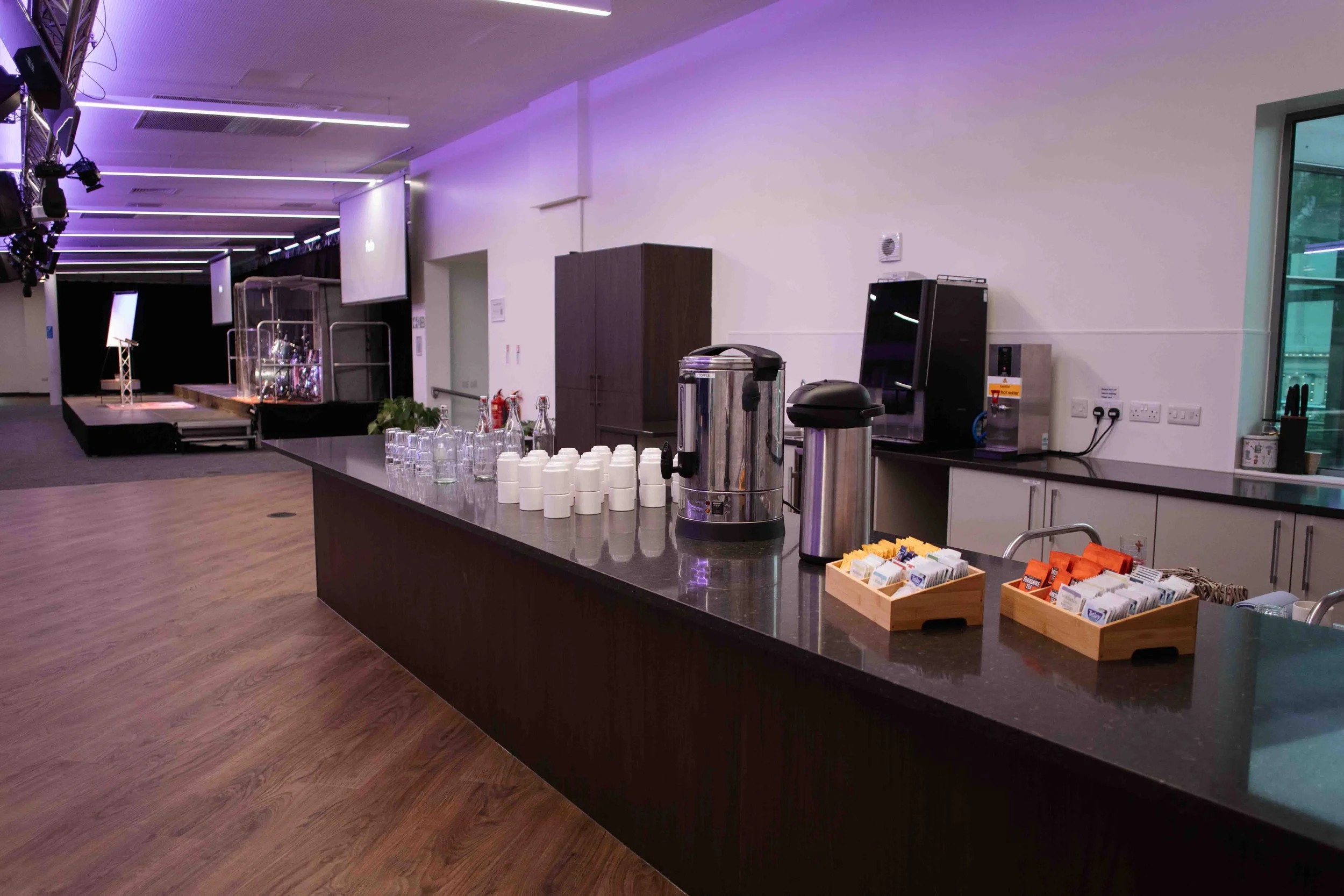 An indoor event space in Bradford with a cafeteria-style serving area, coffee machines, and supply stations on a black counter, with a stage setup in the background illuminated by purple and blue lights.