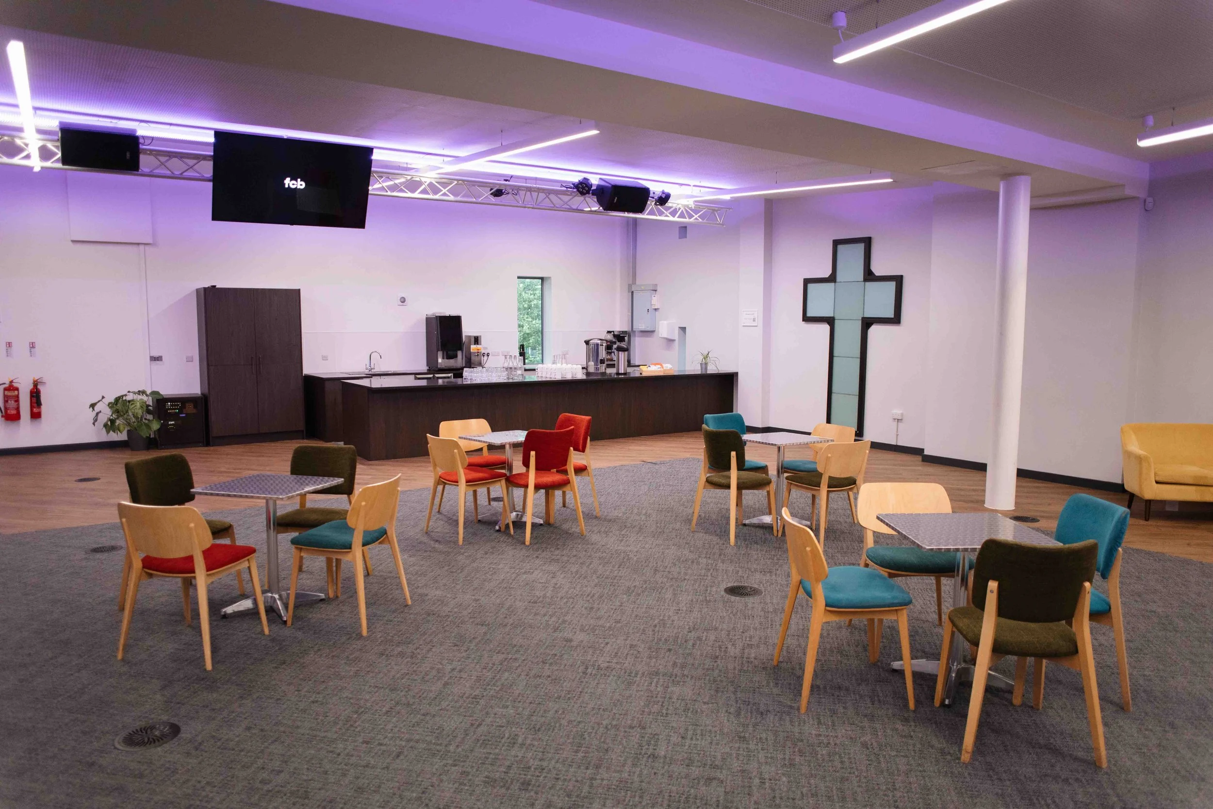 An indoor lounge area with colorful chairs around small tables, a kitchenette with a coffee machine, a potted plant, and a large black cross on the wall. The ceiling has modern lighting and a TV screen displaying 'fcb'.