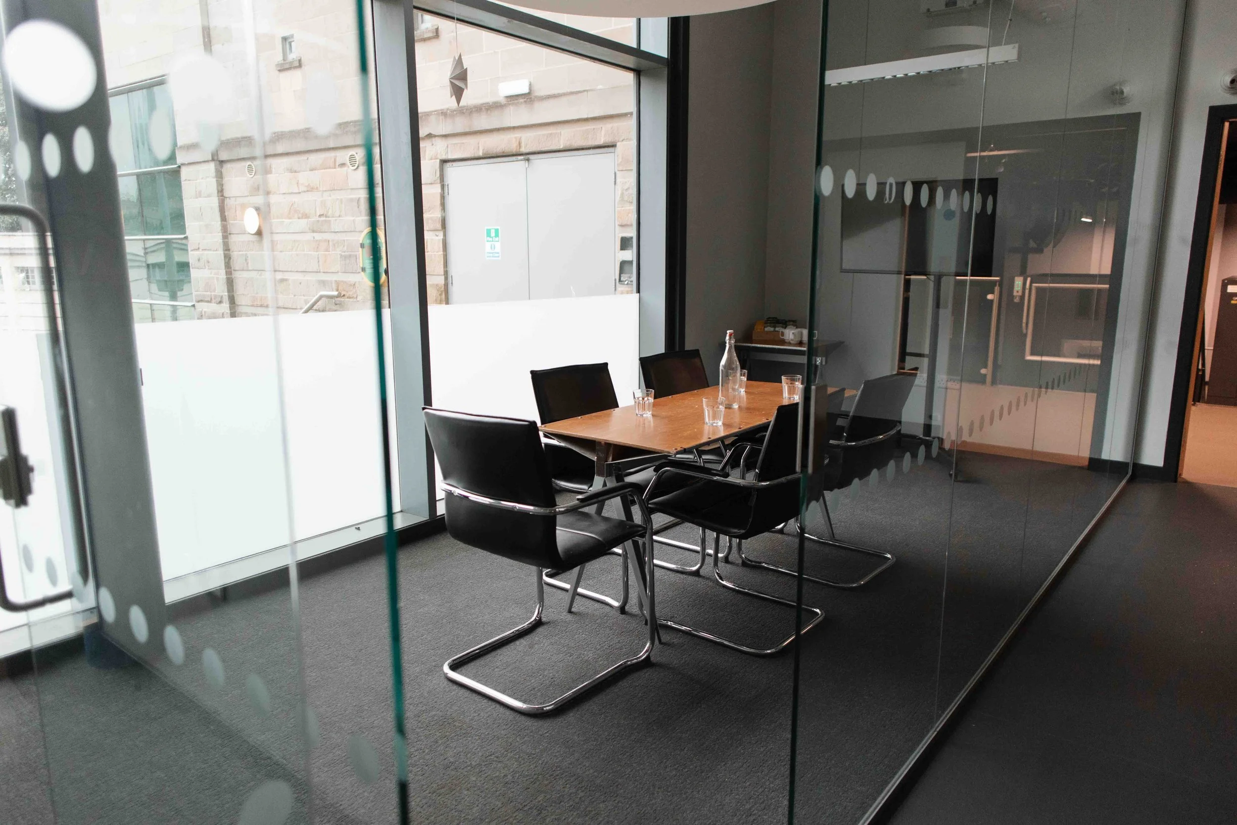 Bradford meeting room with a wooden table, six black chairs, a glass bottle, and glasses. Glass walls, view of an outside stone wall, fire safety sign, and geometric hanging decoration.