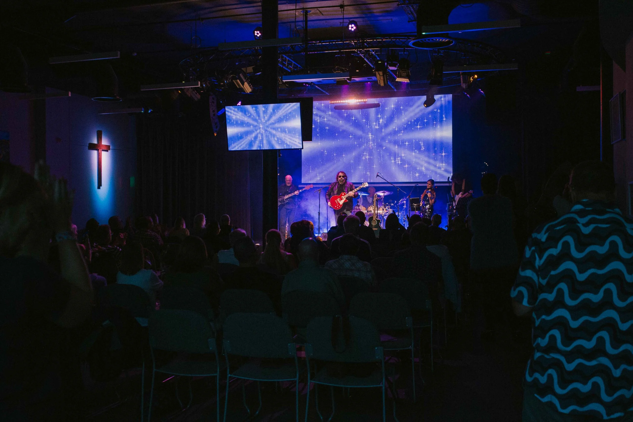 A live music performance in a church with a lit cross on the left wall, a band on stage with guitars and drums, and audience members seated facing the stage.