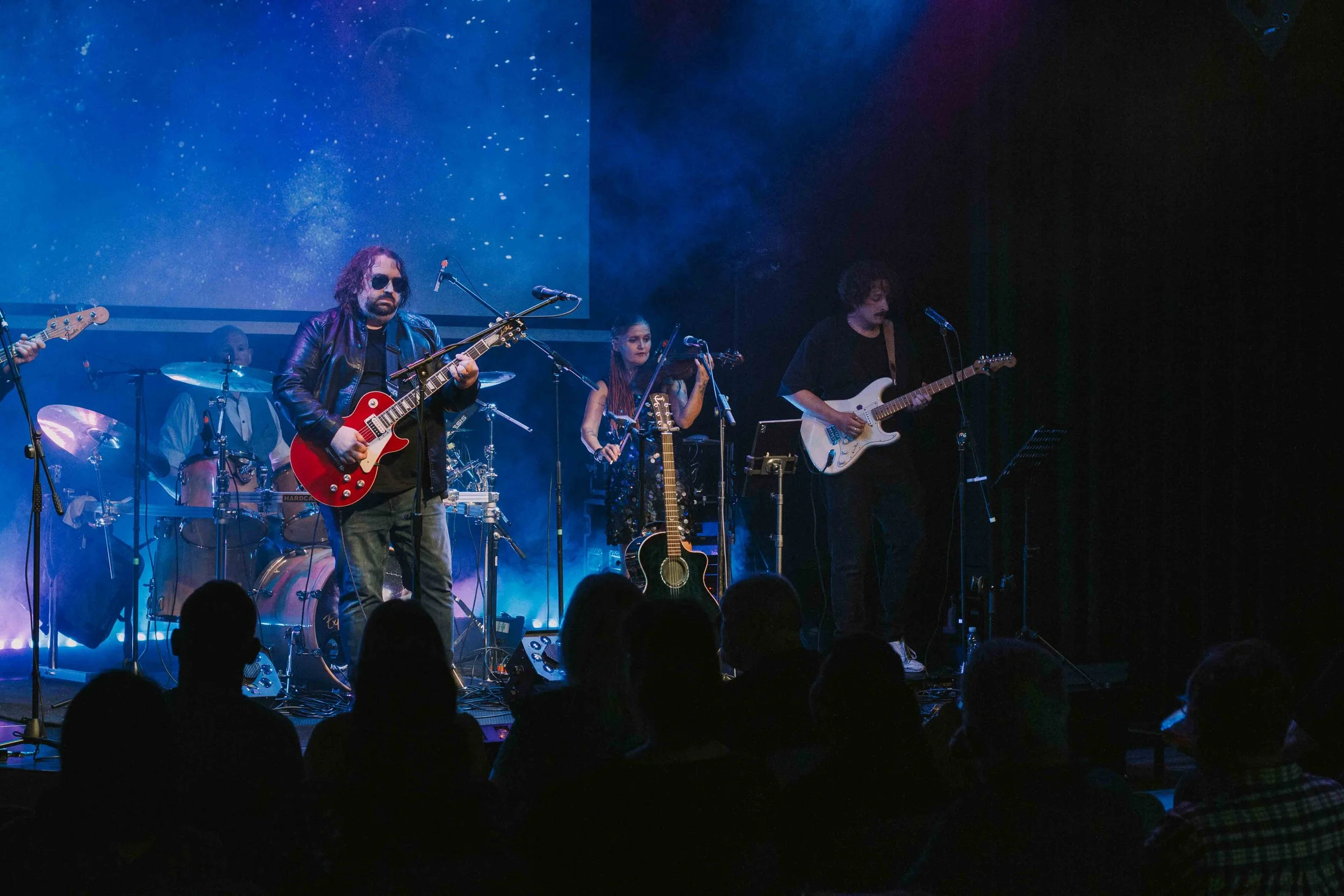A band performing on stage with multiple musicians playing guitars and drums, dark background with blue lighting and audience watching.