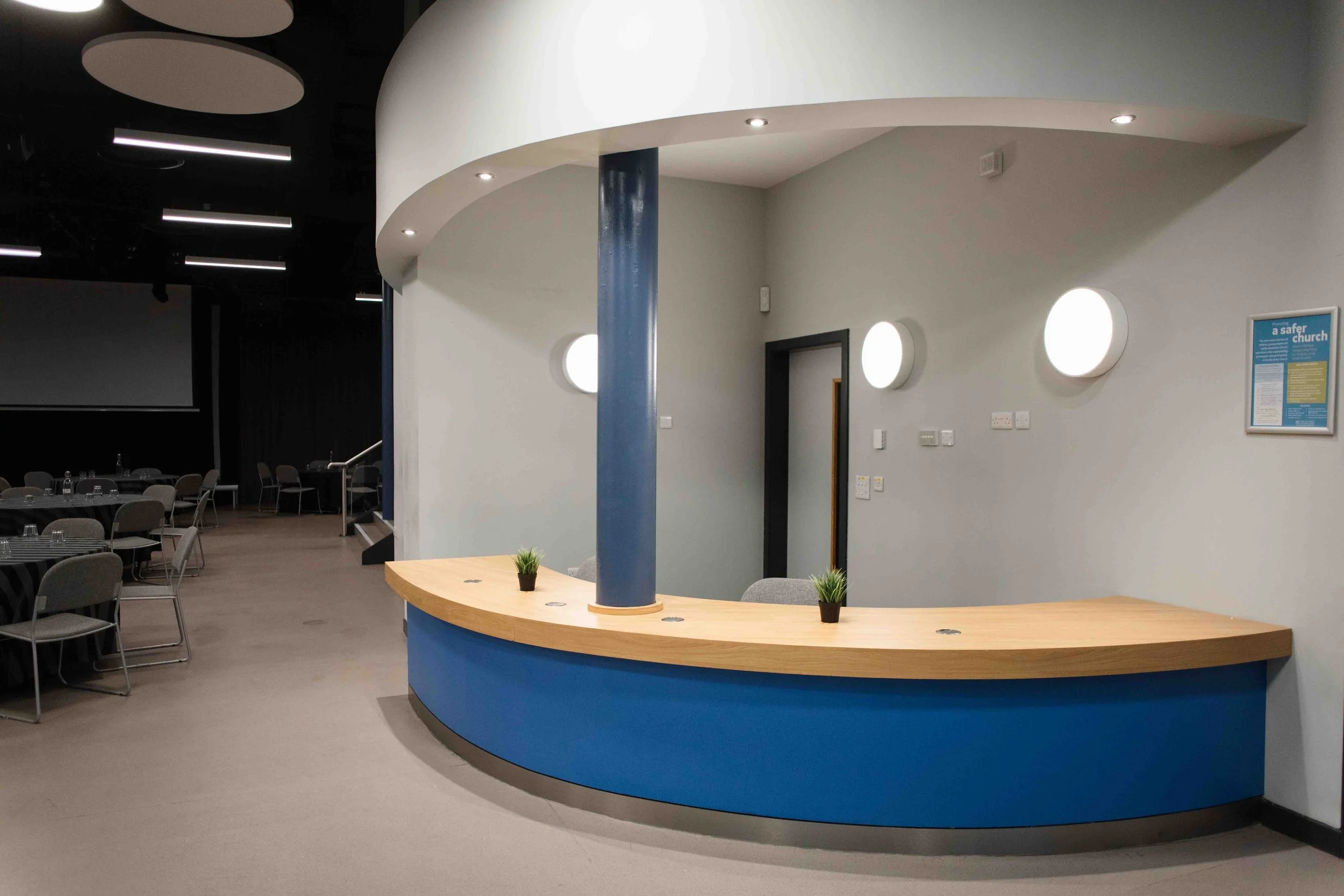 Curved reception desk in Fountains Church Bradford, painted blue and topped with light wood, decorated with small potted plants. To the left, there are tables and chairs for dining or meetings, with a large dark screen or projection area visible.