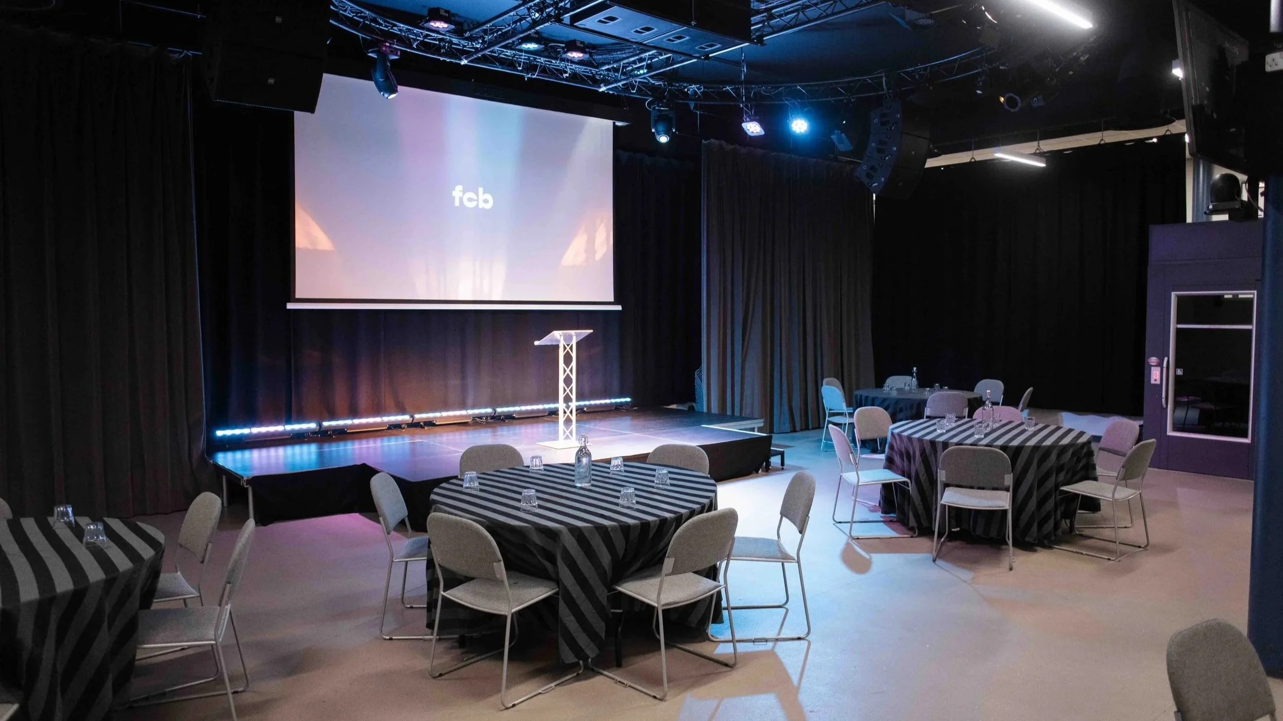 A Bradford event space with tables covered in black and white striped tablecloths, set with water bottles and glasses, facing a stage with a large screen displaying 'fcb' and stage lighting.