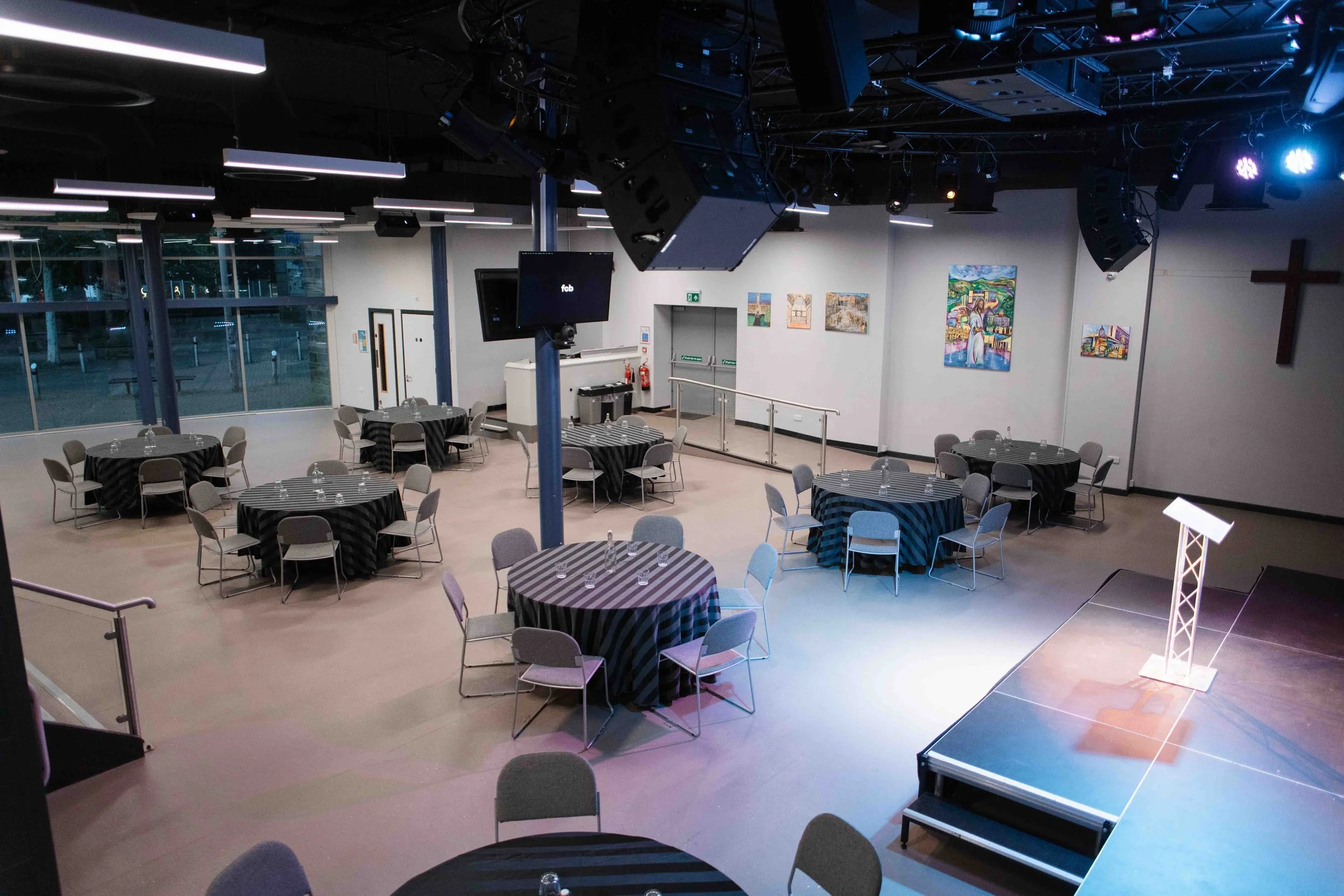 An event space in Bradford with round tables covered with black and white striped tablecloths, each surrounded by gray chairs. There is a small stage in the lower right corner, and a podium with a microphone.