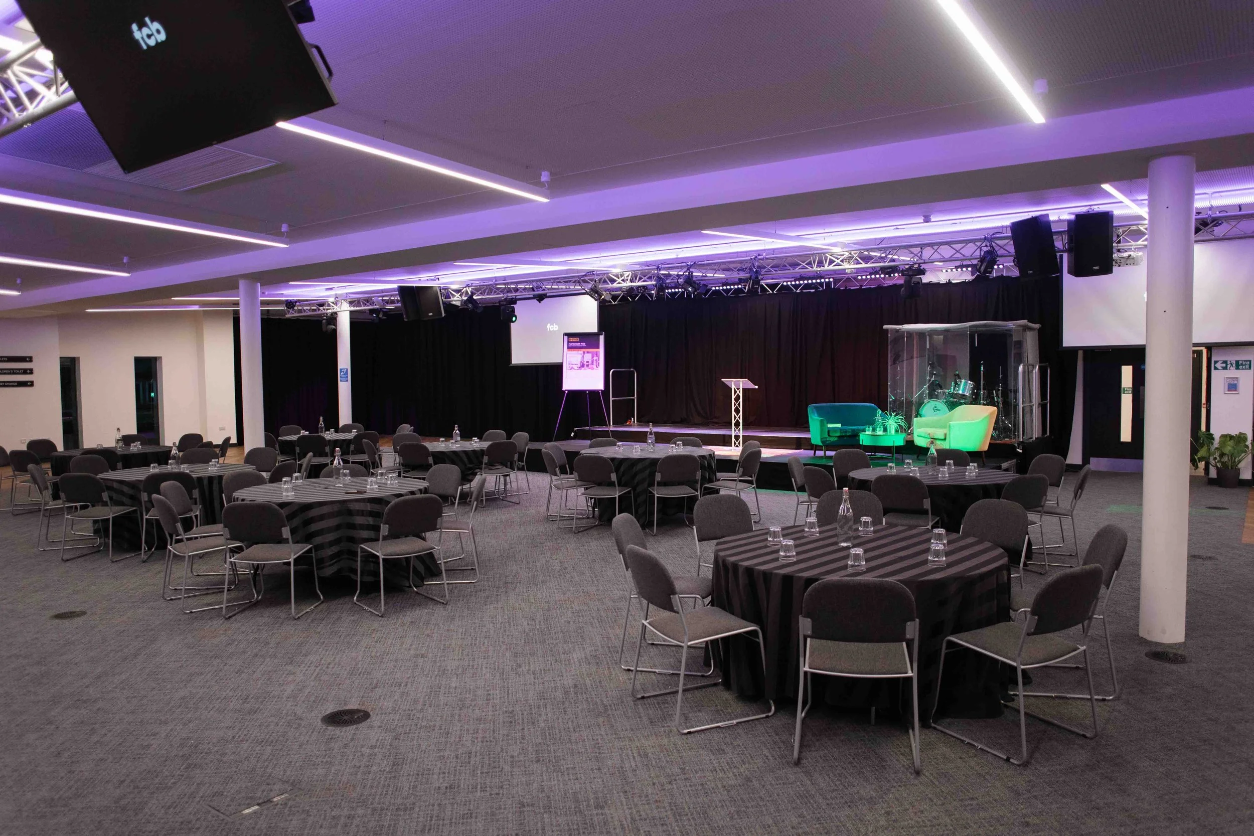 Bradford conference room with round tables covered in black and white striped tablecloths, set with water bottles, chairs, a stage with a lighting truss, two screens, and a seating area with green chairs and drums.