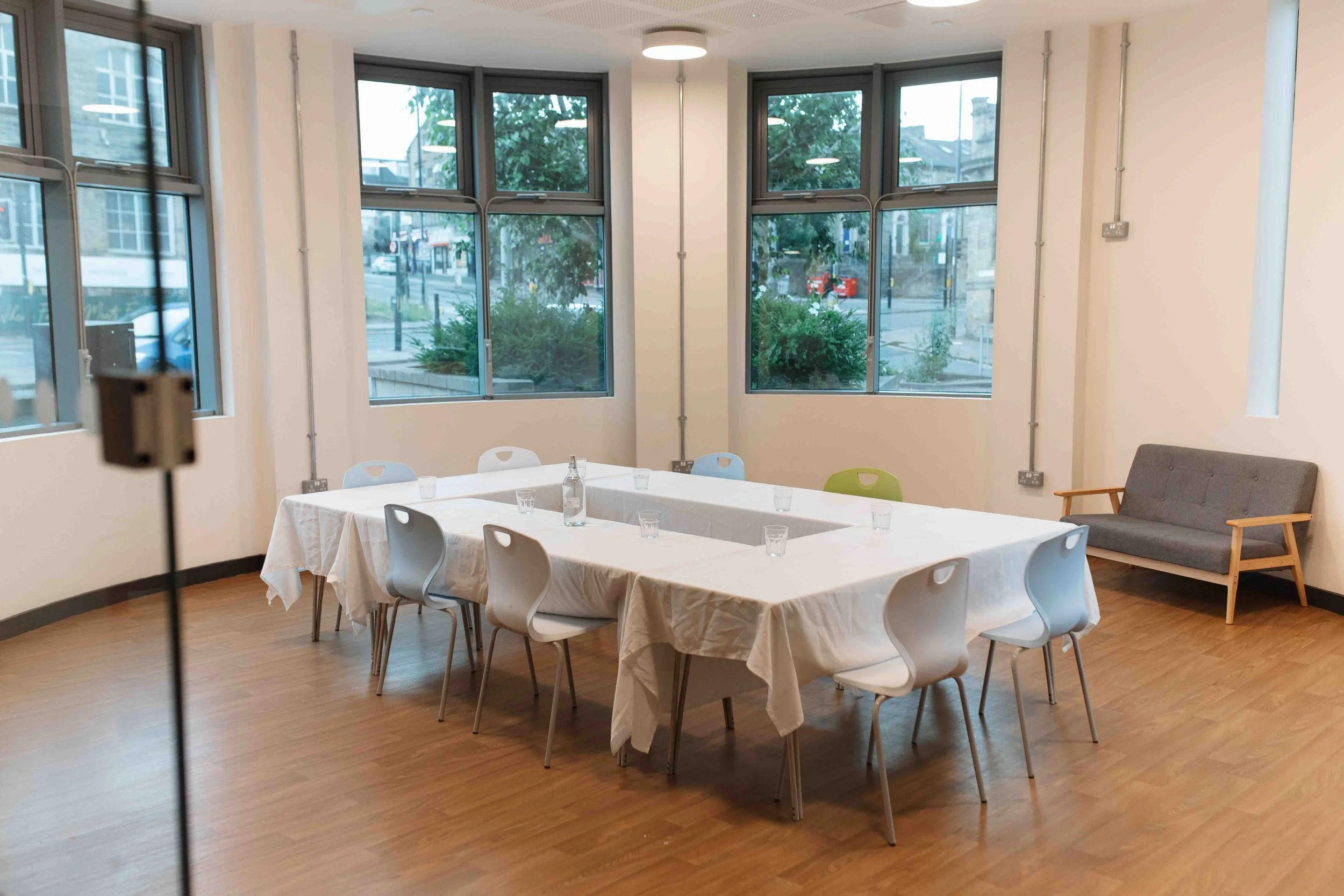 An empty meeting or dining room with a rectangular table covered with a white tablecloth. The table has empty glasses and a small water pitcher. Surrounding the table are white and green chairs. Large windows on the front and side walls offer views o