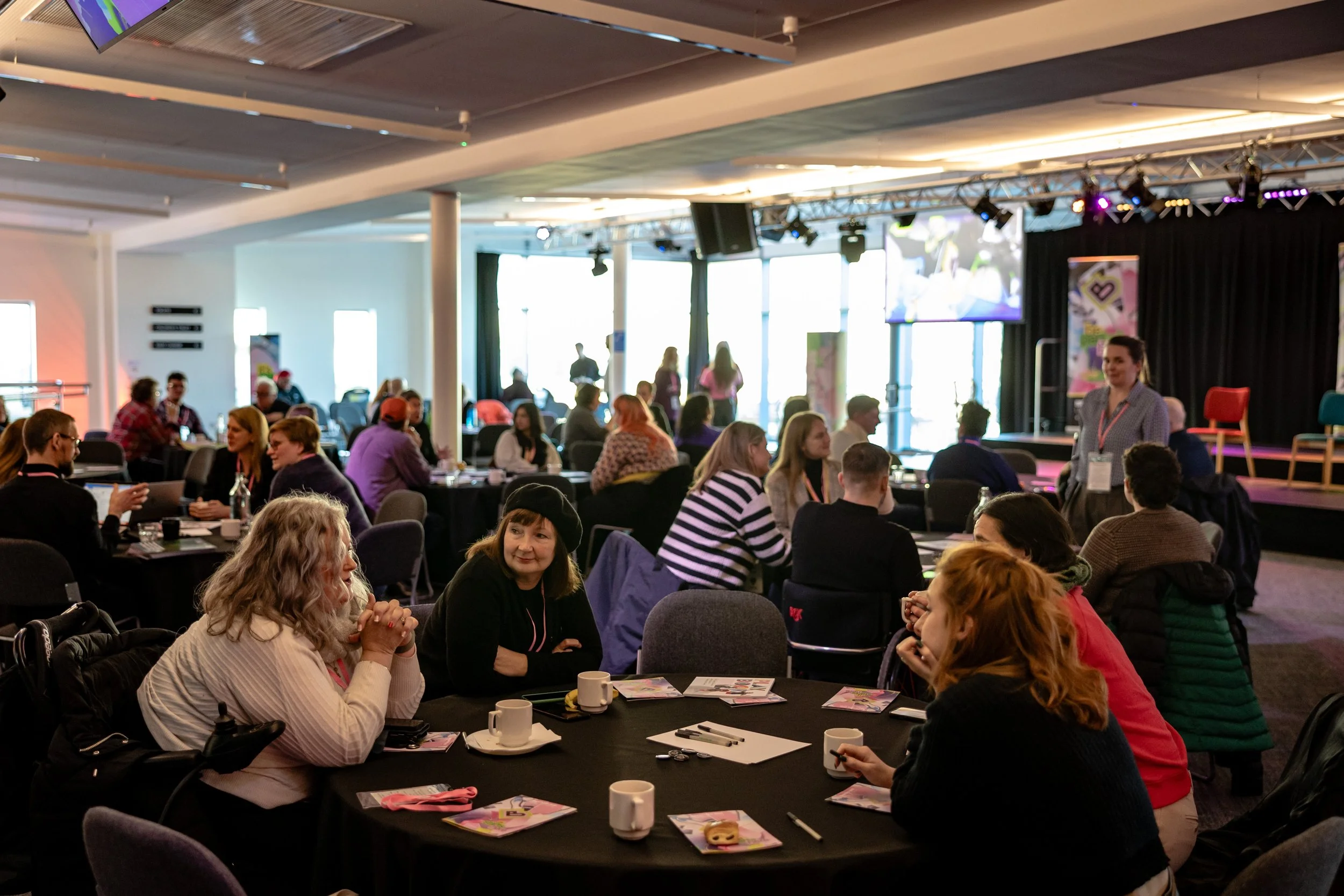 People sitting at round tables in a conference room with a stage and a presenter.