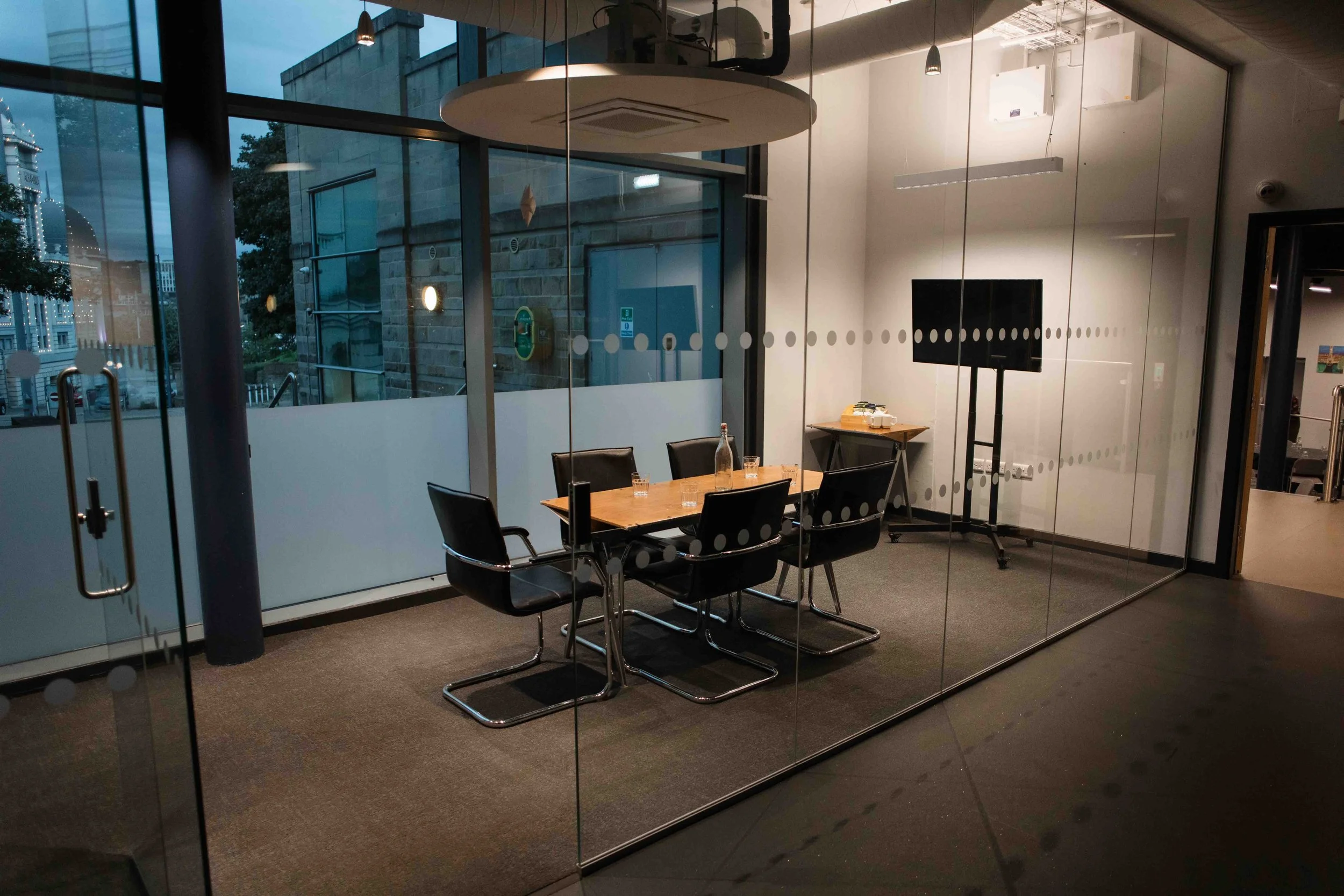 Bradford meeting room with a wooden table, six black chairs, a large display screen, and a glass wall with a dotted pattern. Outside, a city street and buildings are visible during dusk.