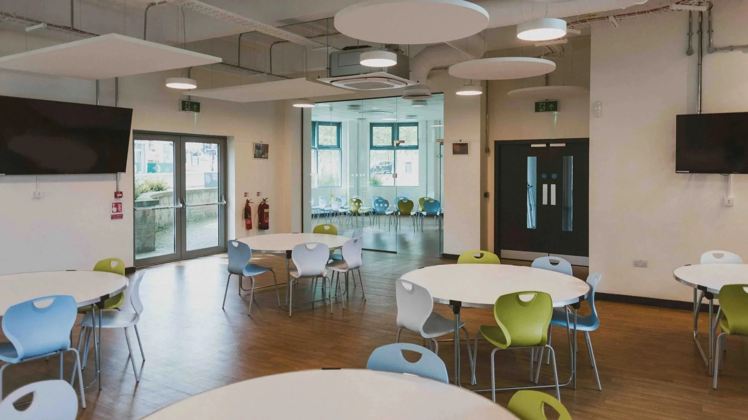 Interior of a modern, brightly lit workshop venue in Bradford with round tables and colourful chairs, an exit door, and a glass-walled room with additional seating