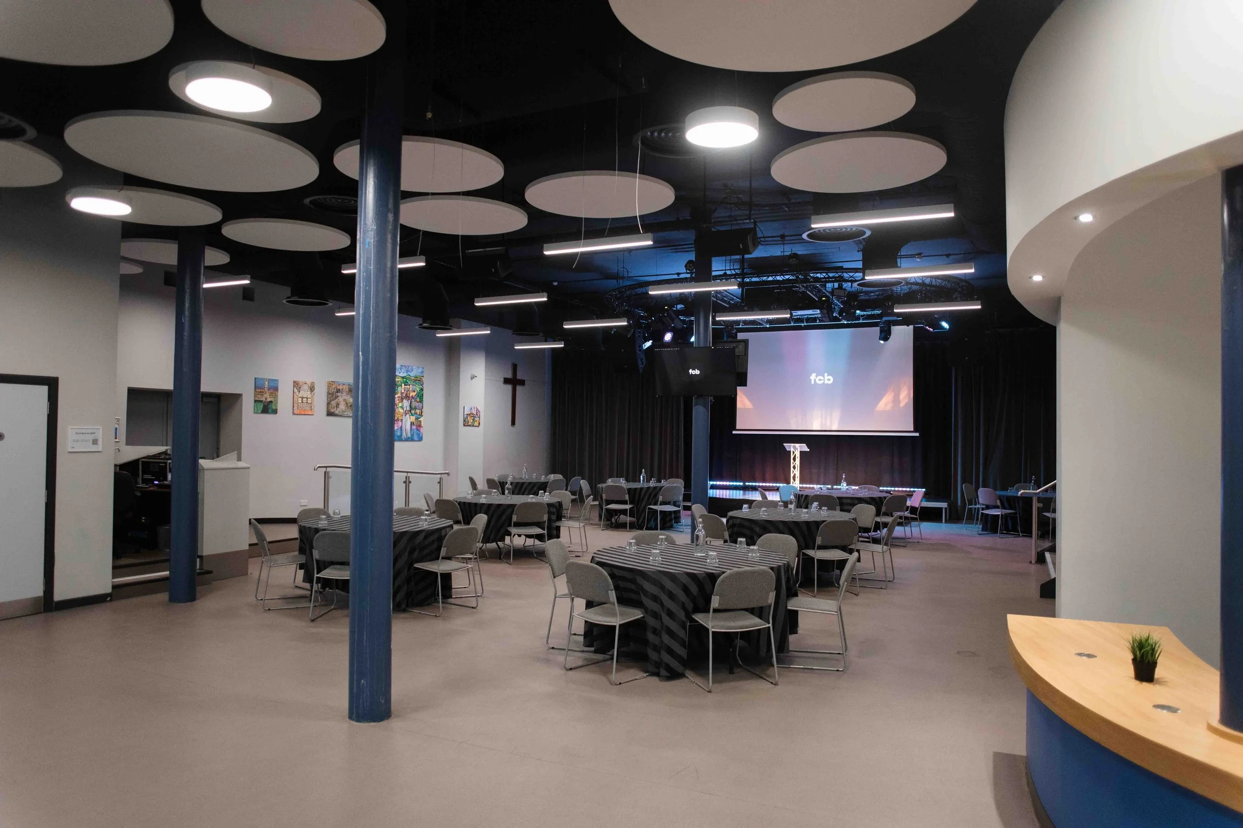 A large indoor event space or conference room with tables and chairs arranged for a gathering or event. The room features modern ceiling design with circular acoustic panels and horizontal light fixtures, a stage with a screen displaying 'fcb,' and s