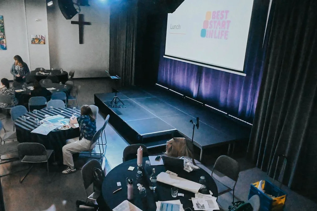 View of a large stage ina. conference room with a large projection screen, surrounded by round tables with conference attendees and their work