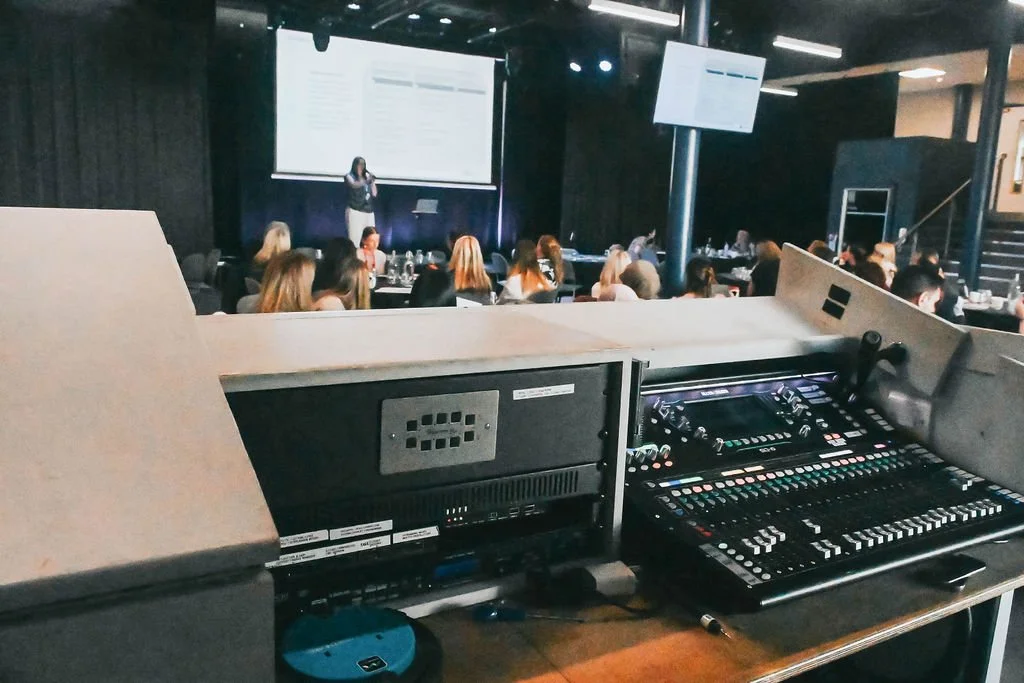 Audio production equipment on a wooden desk, including a digital audio mixer, a computer monitor displaying a digital audio workstation, a keyboard, a mouse, a second monitor, and a Sennheiser speaker, with a window and ceiling light in the backgroun
