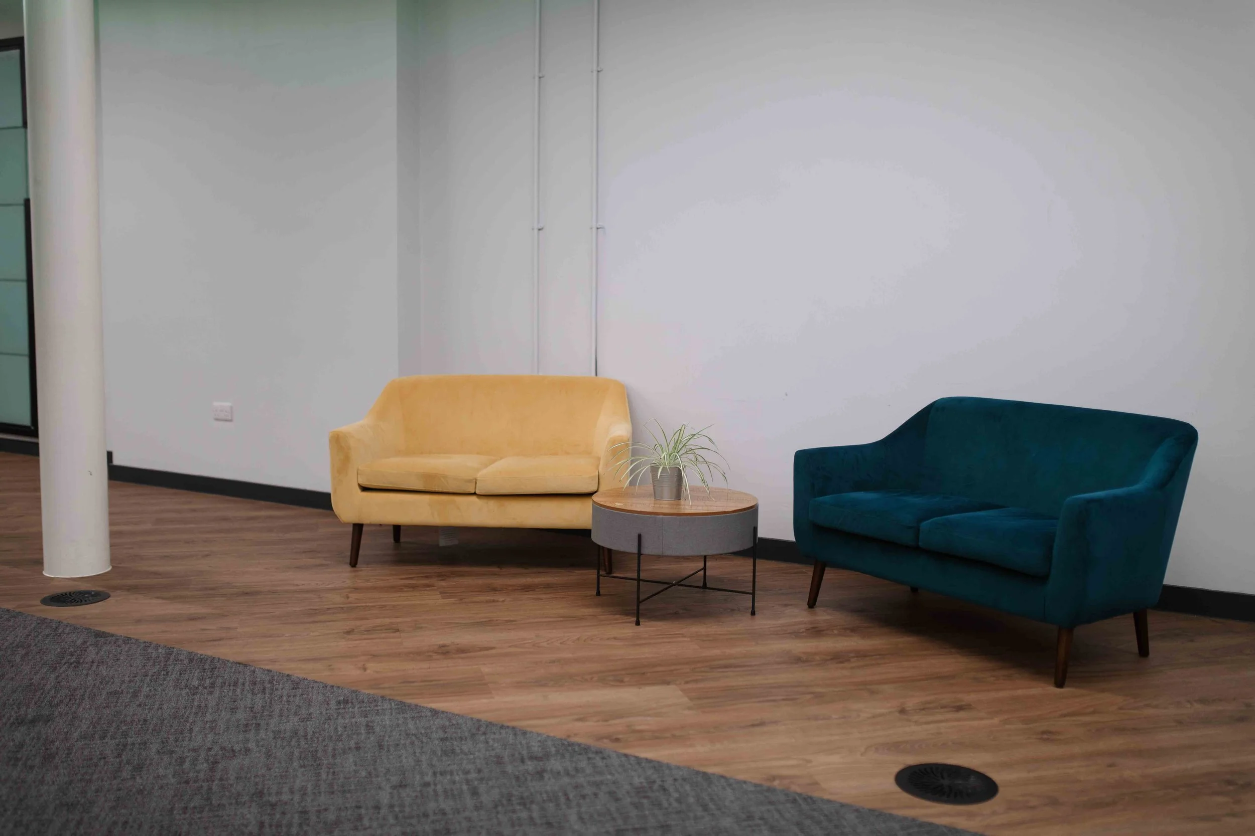 Interior of a modern waiting area with a yellow and green armchair, a small wooden round table with a potted plant, and a section of a teal armchair, with a white wall and wooden flooring.