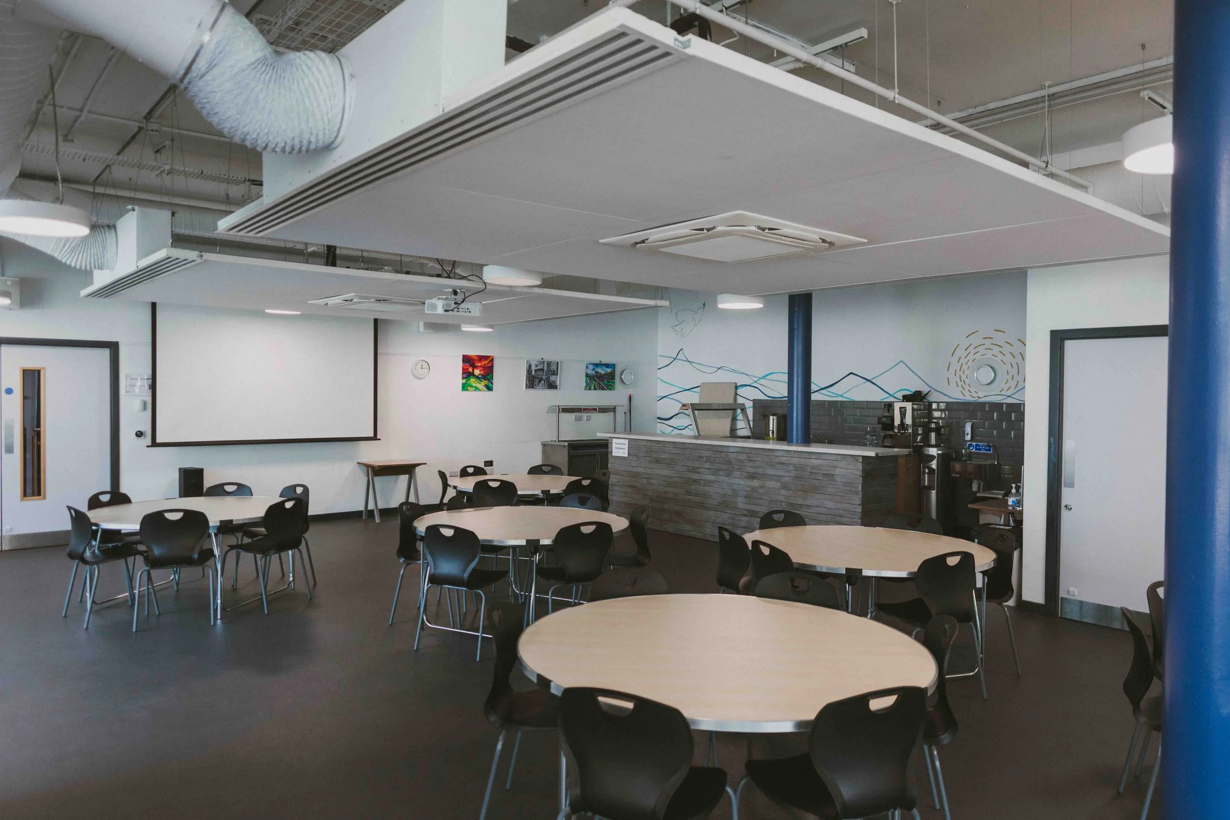 A community room with round tables and black chairs, a wall with some colorful artwork, a whiteboard, a clock, and a counter with coffee machines and supplies. Ceiling has ventilation ducts and lighting fixtures.