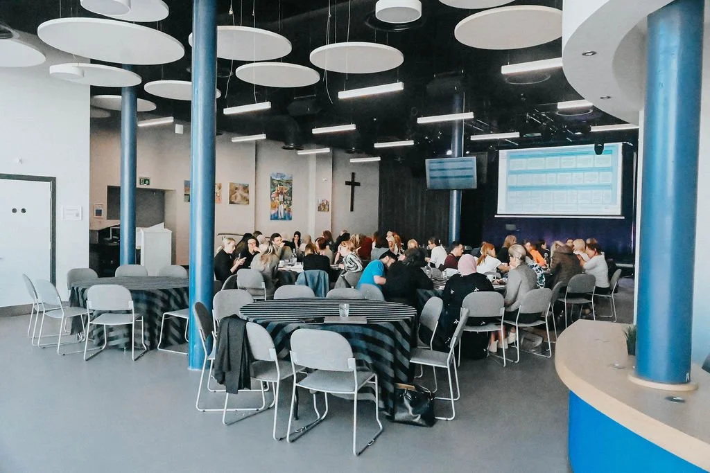 Spacy and well equipped conference space with large round tables and chairs, a stage with a microphone stand and  two large screens.