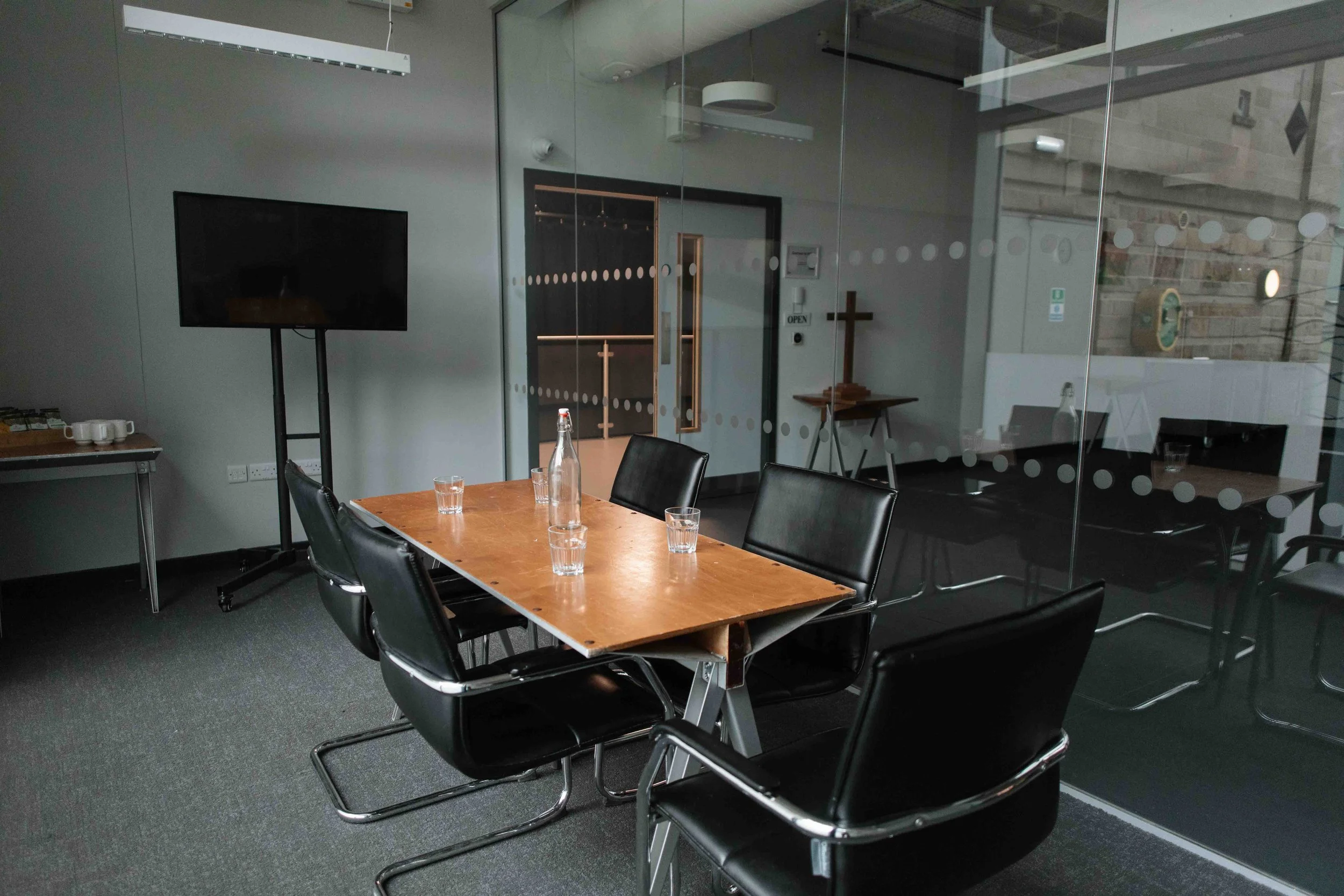 A modern conference room with a wooden table surrounded by black chairs, a glass wall with dotted white patterns, a large flat-screen monitor on a stand, and a glass door with an 'OPEN' sign.