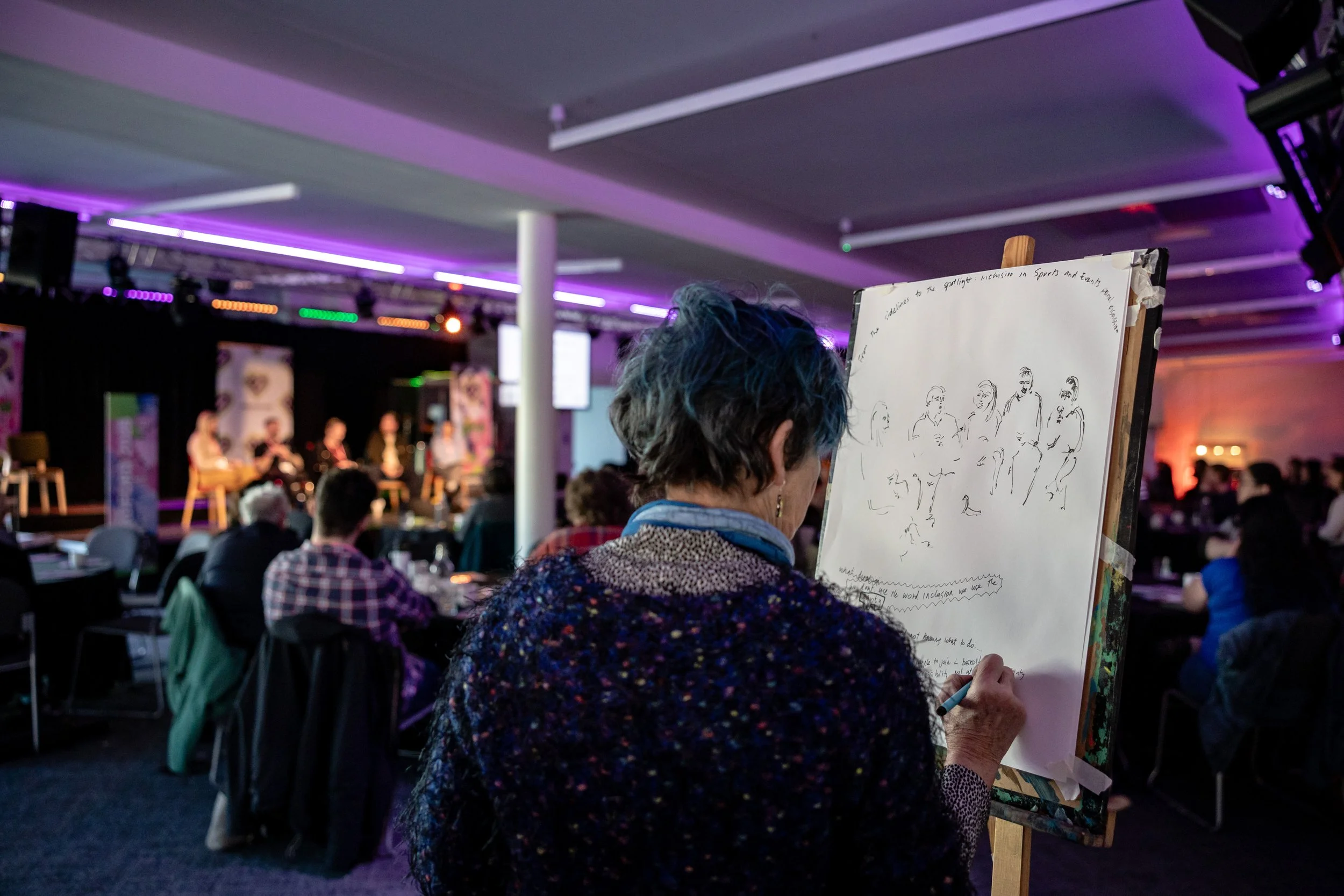 A woman with short blue hair sketches on a large sheet of paper, seated at an event with a panel of speakers on stage in the background. The audience is seated at tables, and colorful stage lights illuminate the room.