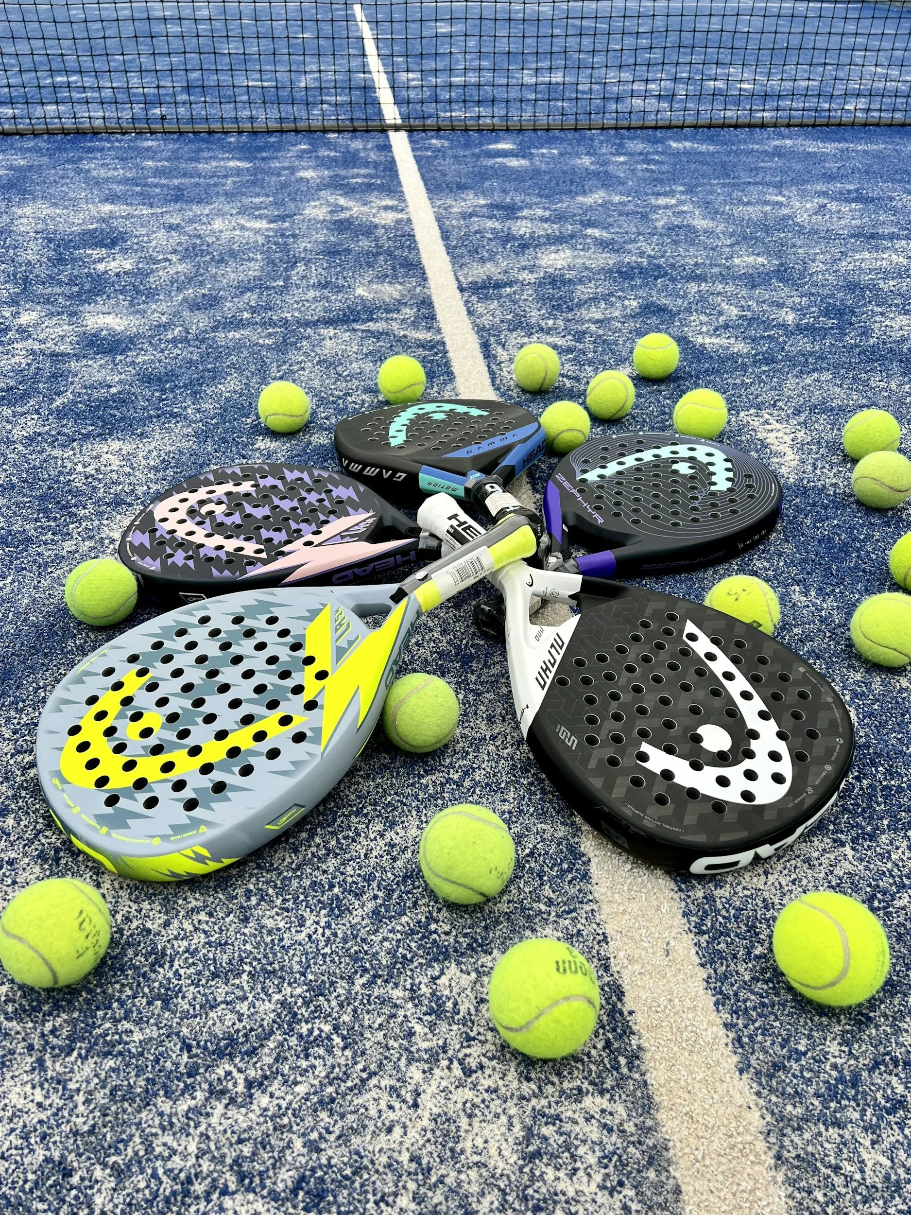 A collection of tennis rackets and tennis balls scattered on a blue tennis court with a net in the background.