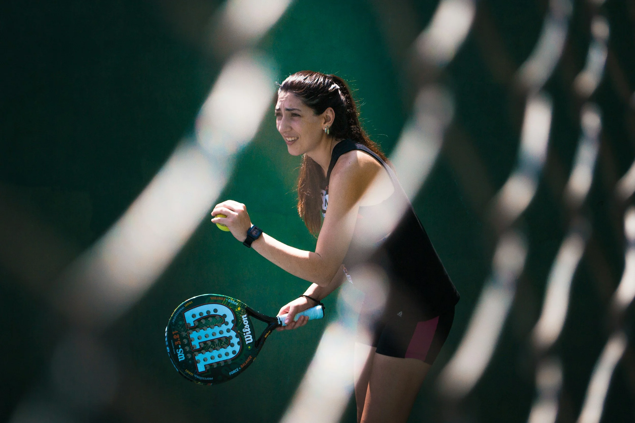 A female tennis player with long dark hair, wearing a black tank top, holding a tennis racket, and smiling as she prepares to serve, seen through a chain-link fence.