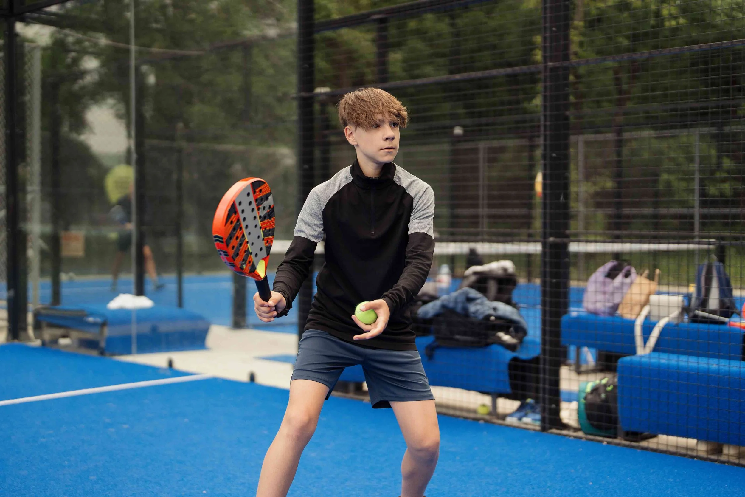 A young boy with short red hair wearing a black and gray long sleeve athletic shirt and dark shorts holding a tennis racket and a tennis ball at an outdoor tennis court.